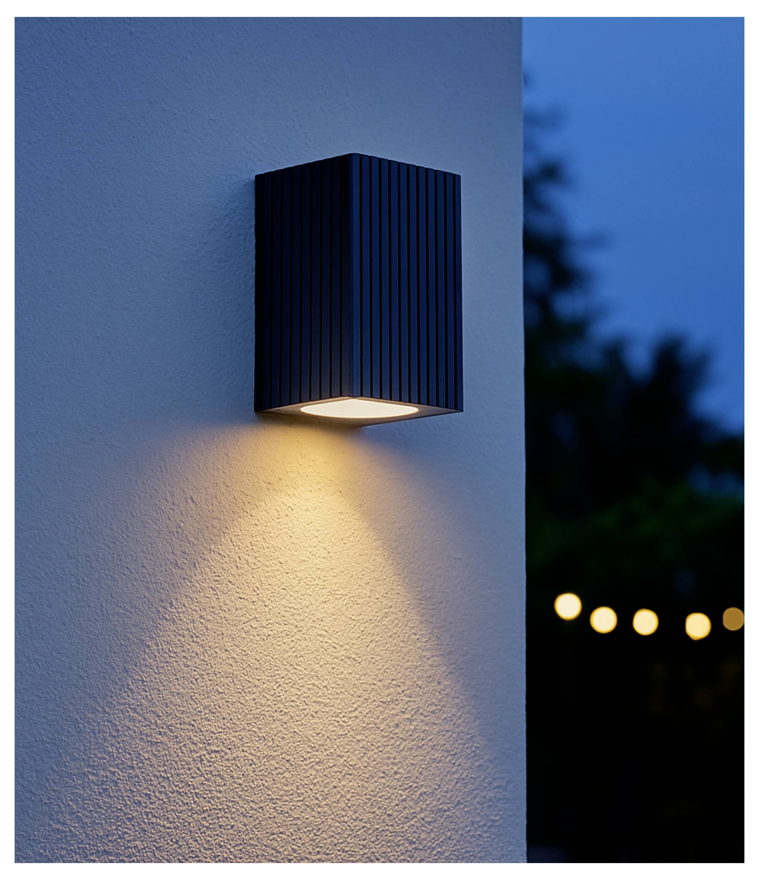 Zwarte buitenwandlamp werpt een warme lichtkegelop een gestructureerde stucowand in de schemering, met onscherpe tuinlichten op de achtergrond.
