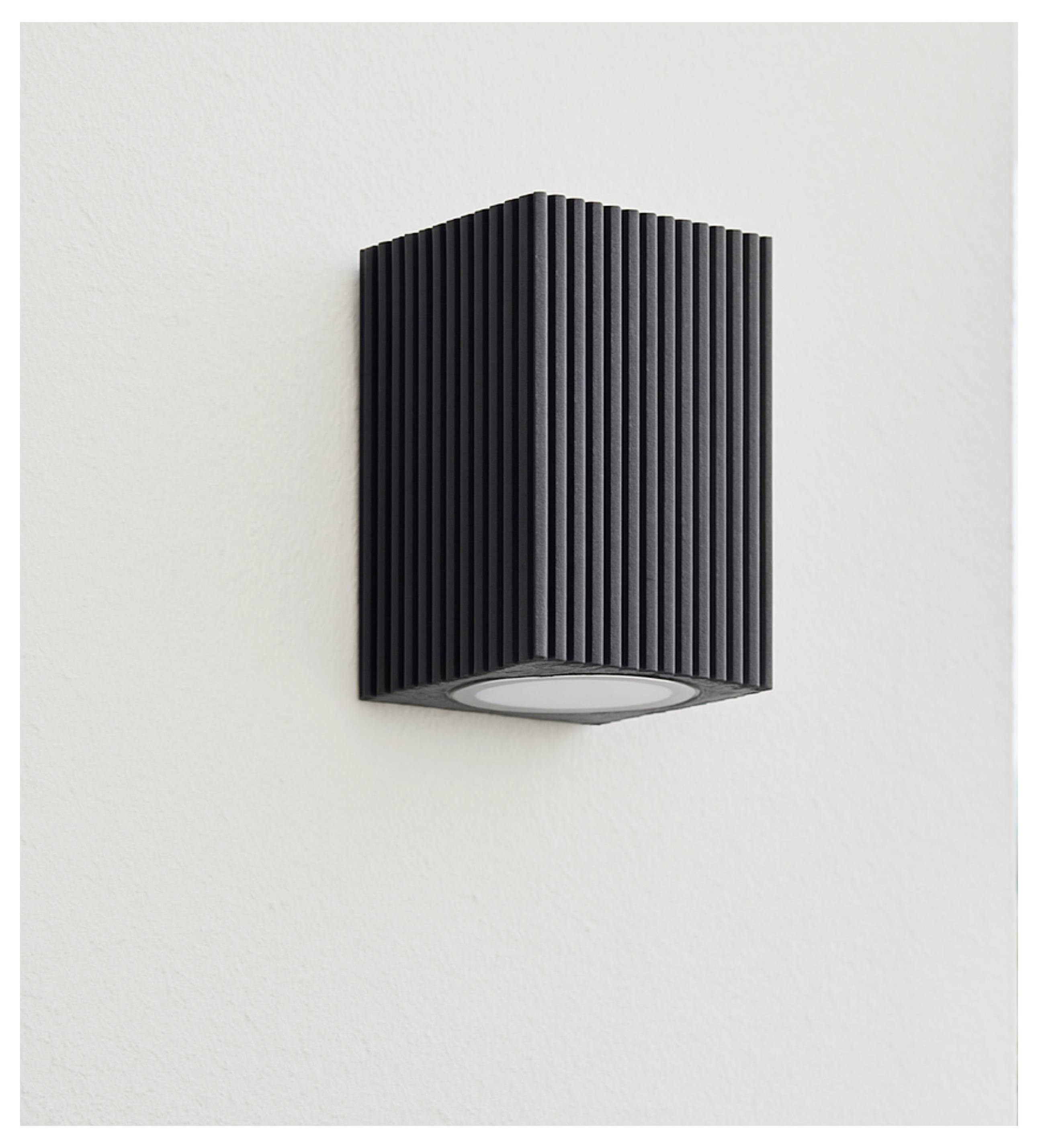 Zwarte geribde rechthoekige wandlamp gemonteerd op een lichte muur, met een ovale naar beneden gerichte lichtopening.