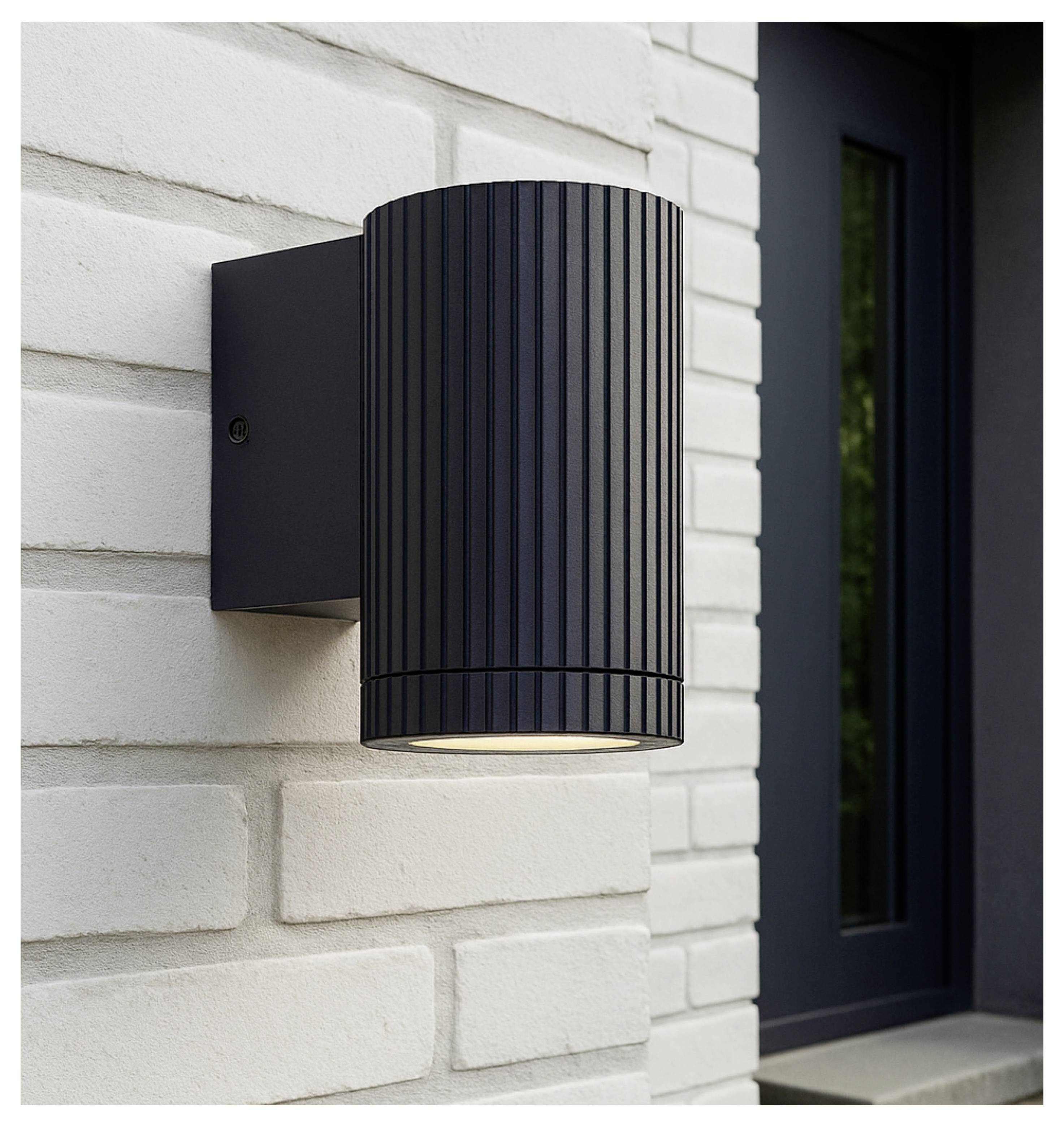 Zwarte cilindrische wandlamp gemonteerd op een lichte bakstenen buitengevel naast een deuropening, van onderaf verlicht.