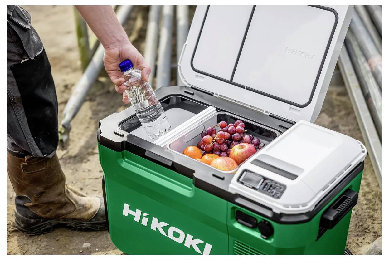 Een werker op een bouwplaats zet een waterfles in een open groene HiKOKI koelbox op wielen naast gestapelde metalen buizen.