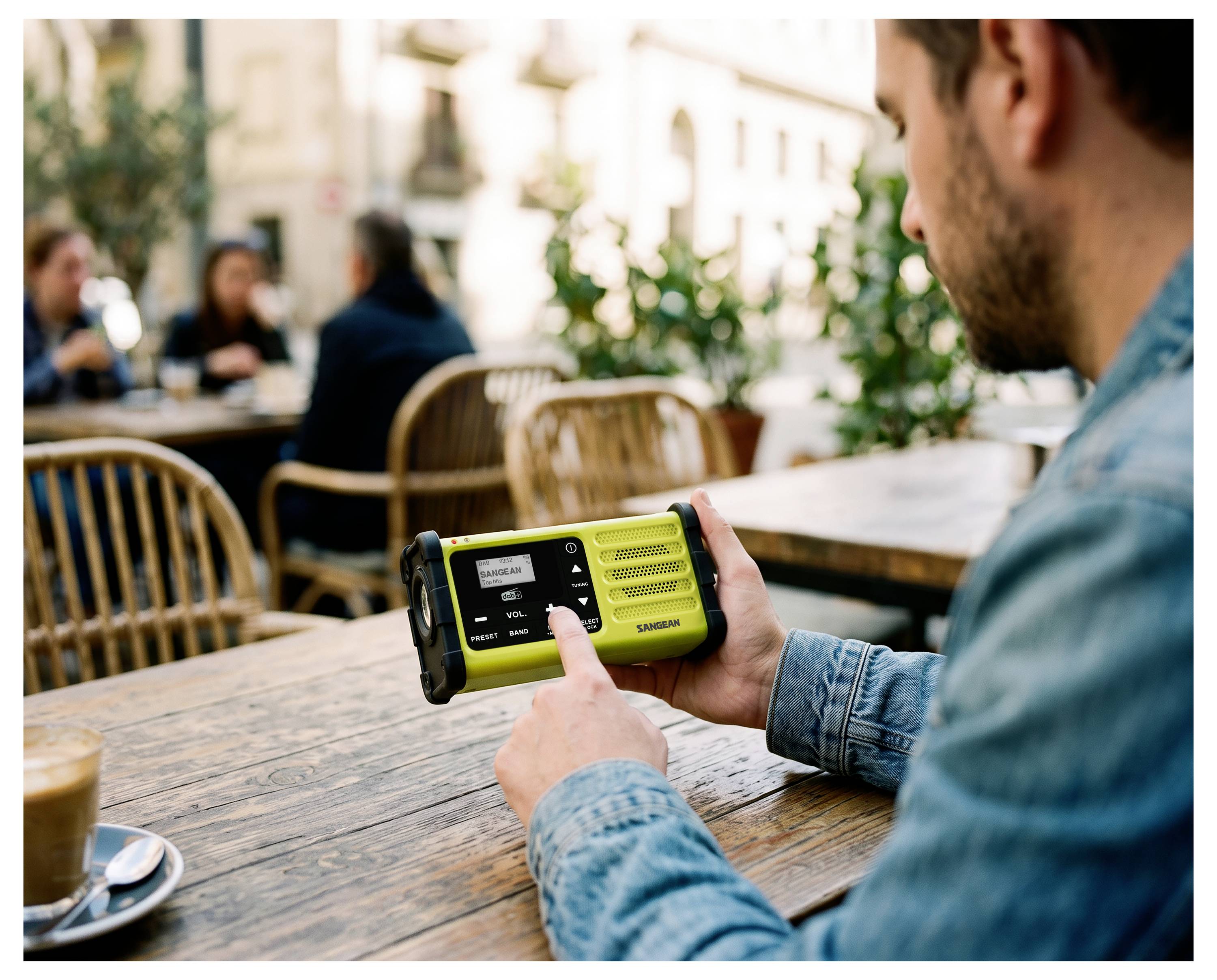 Man die een handheld geel-zwart radio- of sprekekapparaat gebruikt aan een tafel op een buitencafé, met onscherp zichtbare zittende personen op de achtergrond.