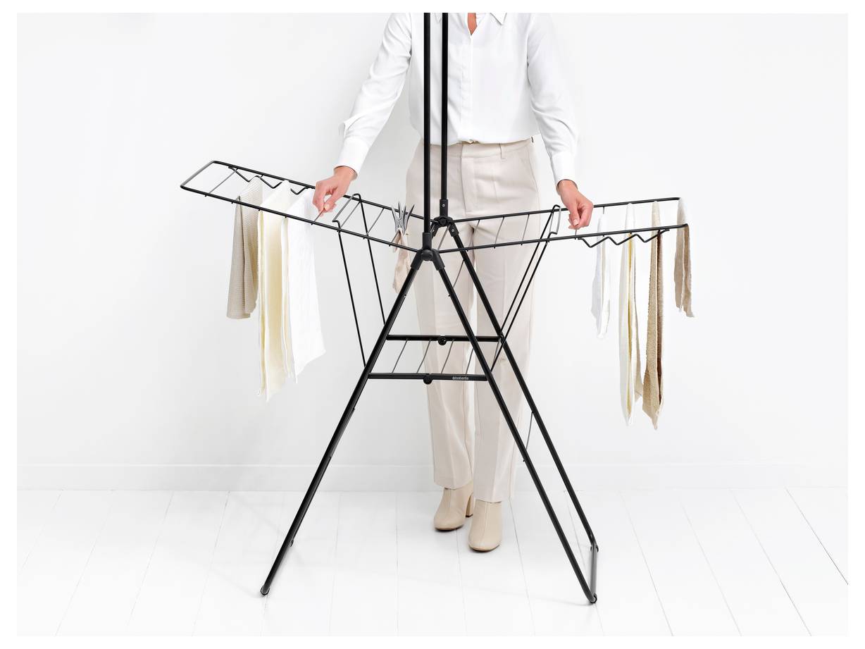 Een persoon in witte kleding staat achter een vrijstaand droogrek of kledingrek en arrangeert opgehangen kledingstukken.