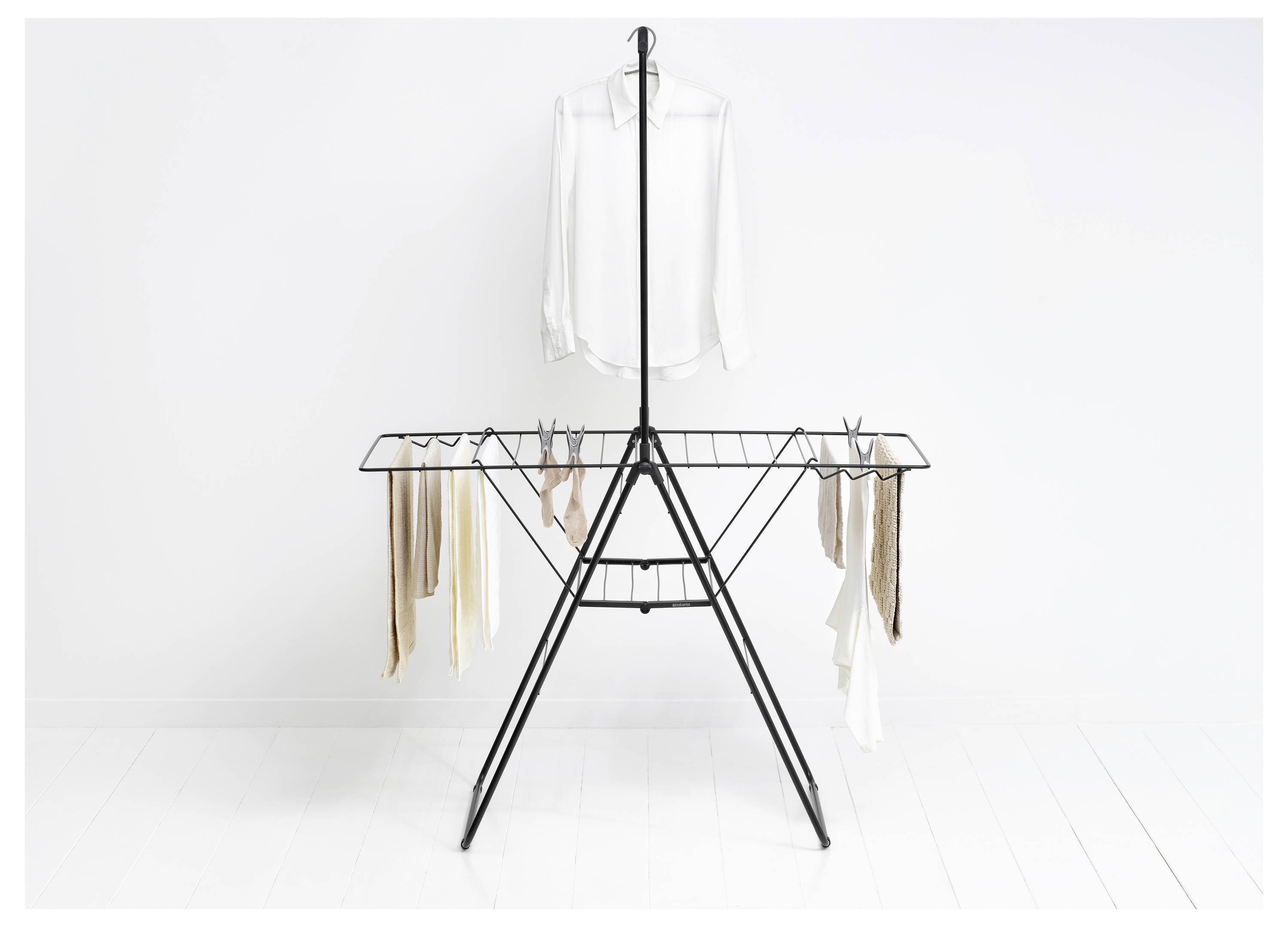 Een zwart droogrek houdt een wit shirt en enkele lichtgekleurde kledingstukken in een minimale witte kamer.