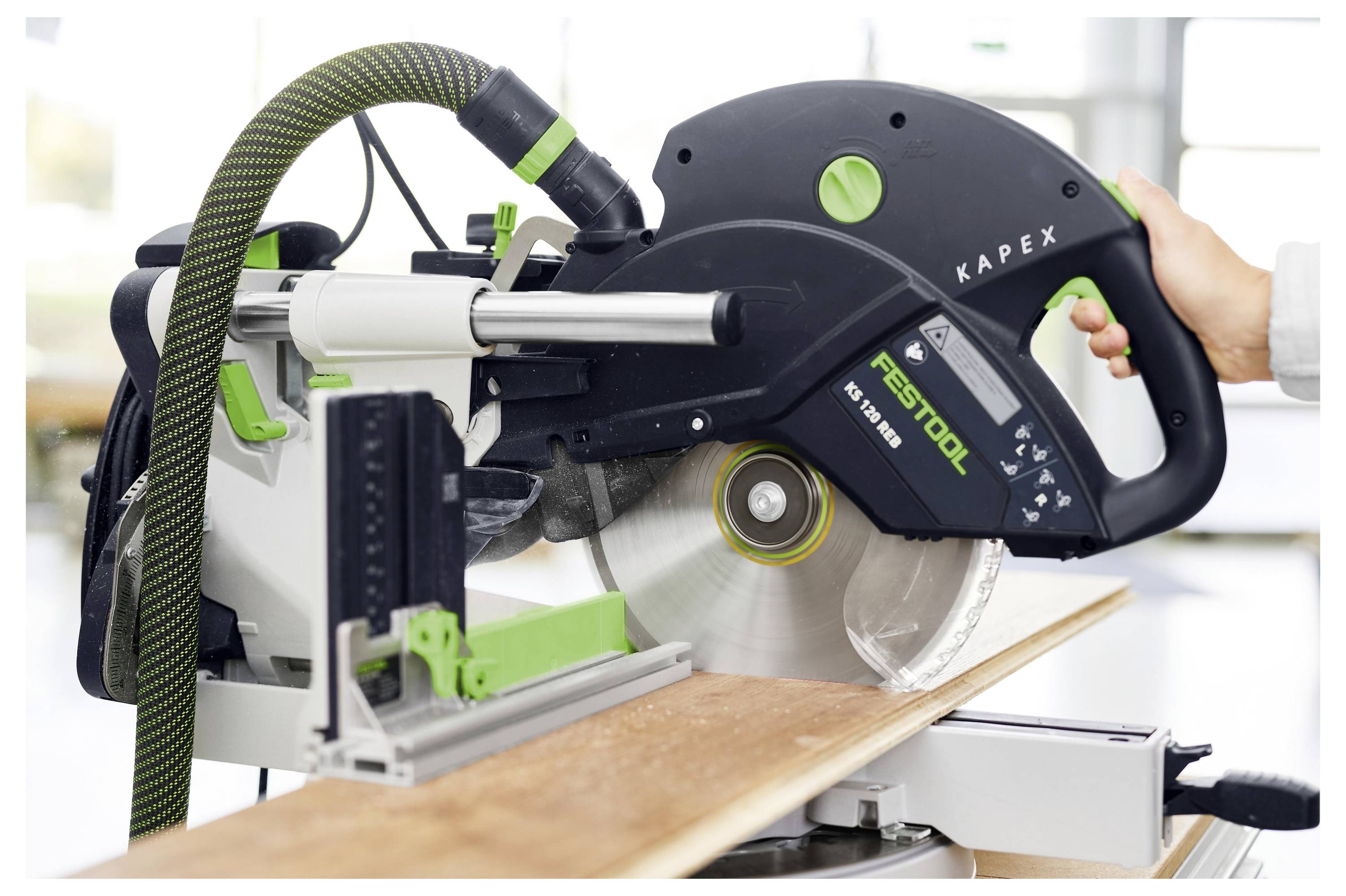 Een Festool Kapex schuifvergrendeling verstekzaag snijdt een houten plank, met een hand die de handgreep vasthoudt en het zaagblad dat draait in een werkplaats.