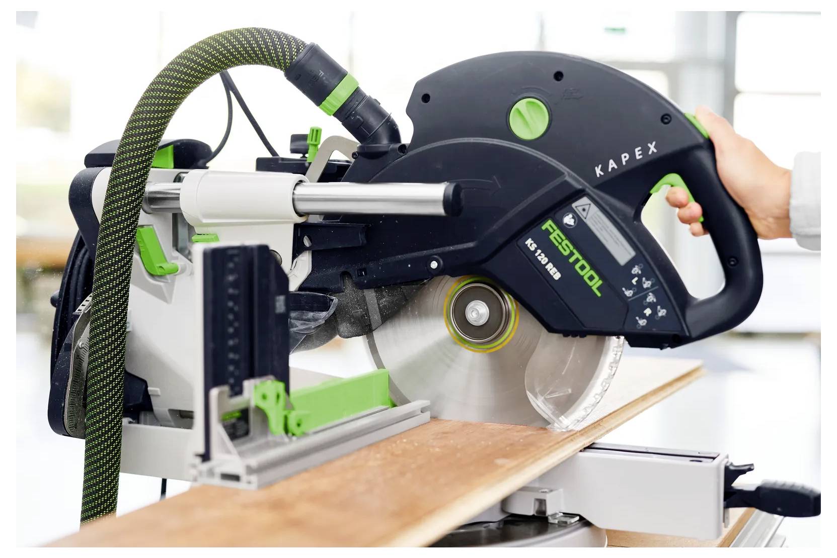 Een Festool Kapex schuifmergel zag die een houten plank zaagt, met een hand die het handvat vasthoudt en het blad blootgesteld in gebruik.