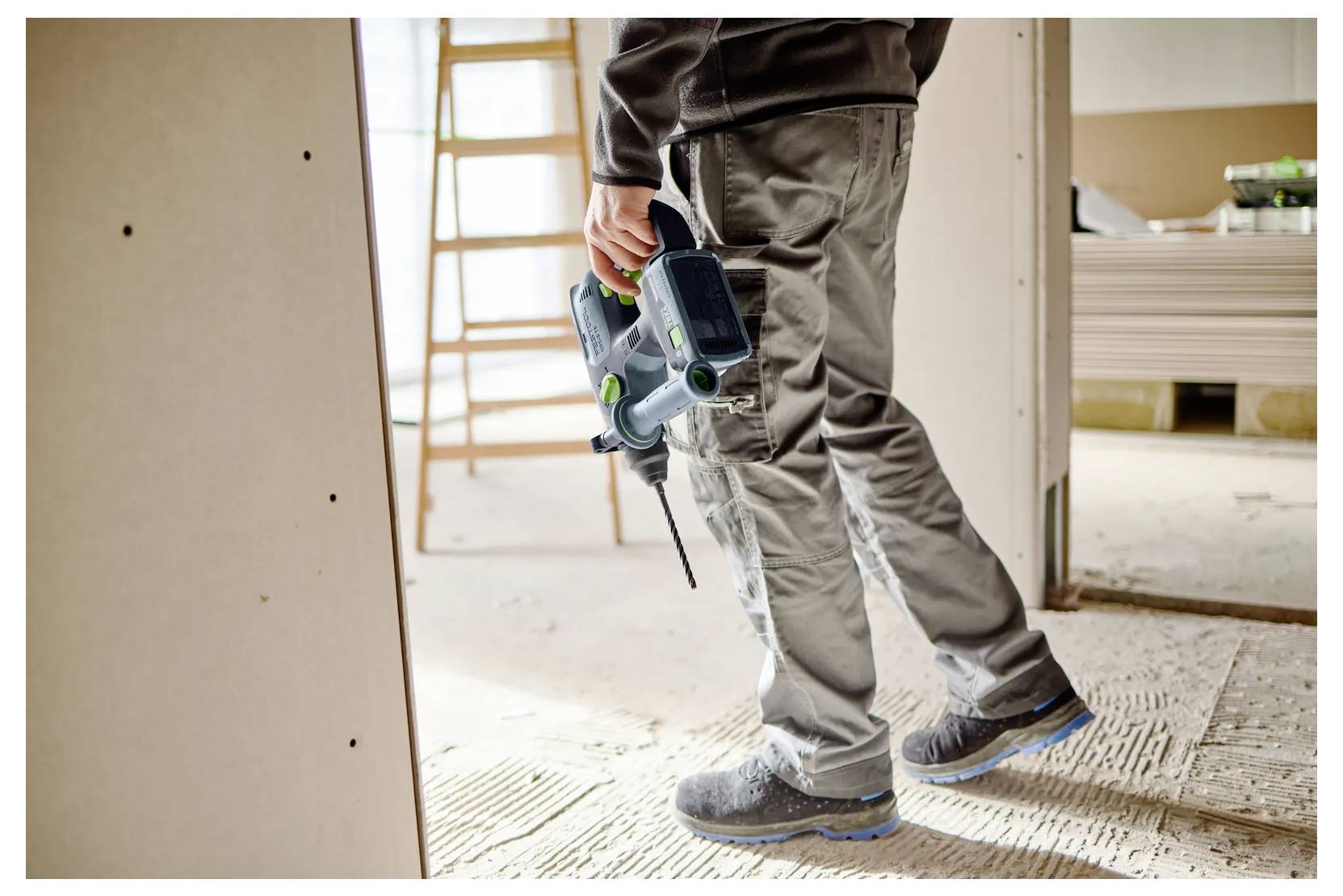 Bouwvakker met een elektrische boor op een renovatielocatie, staand op onafgewerkte vloering met ladders en gipsplaten op de achtergrond.