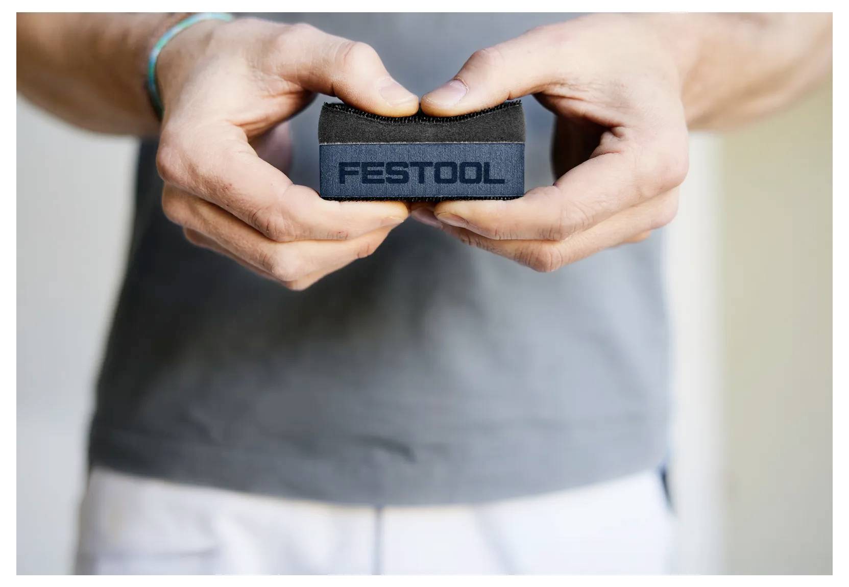 Handen die een kleine zwarte stofband met het label 'FESTOOL' tegen een grijze shirtachtergrond houden.