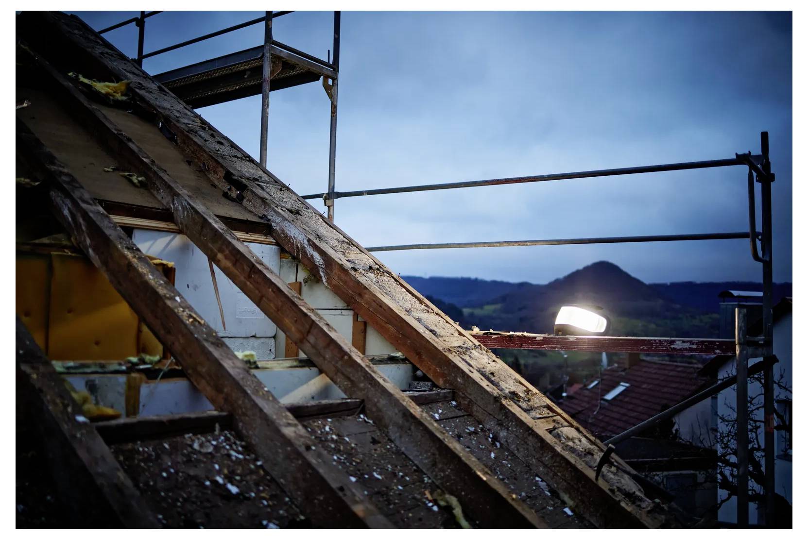 Verslechterende houten dakbalken onder steigers bij schemering, met een felle werklamp en verre bergen op de achtergrond.