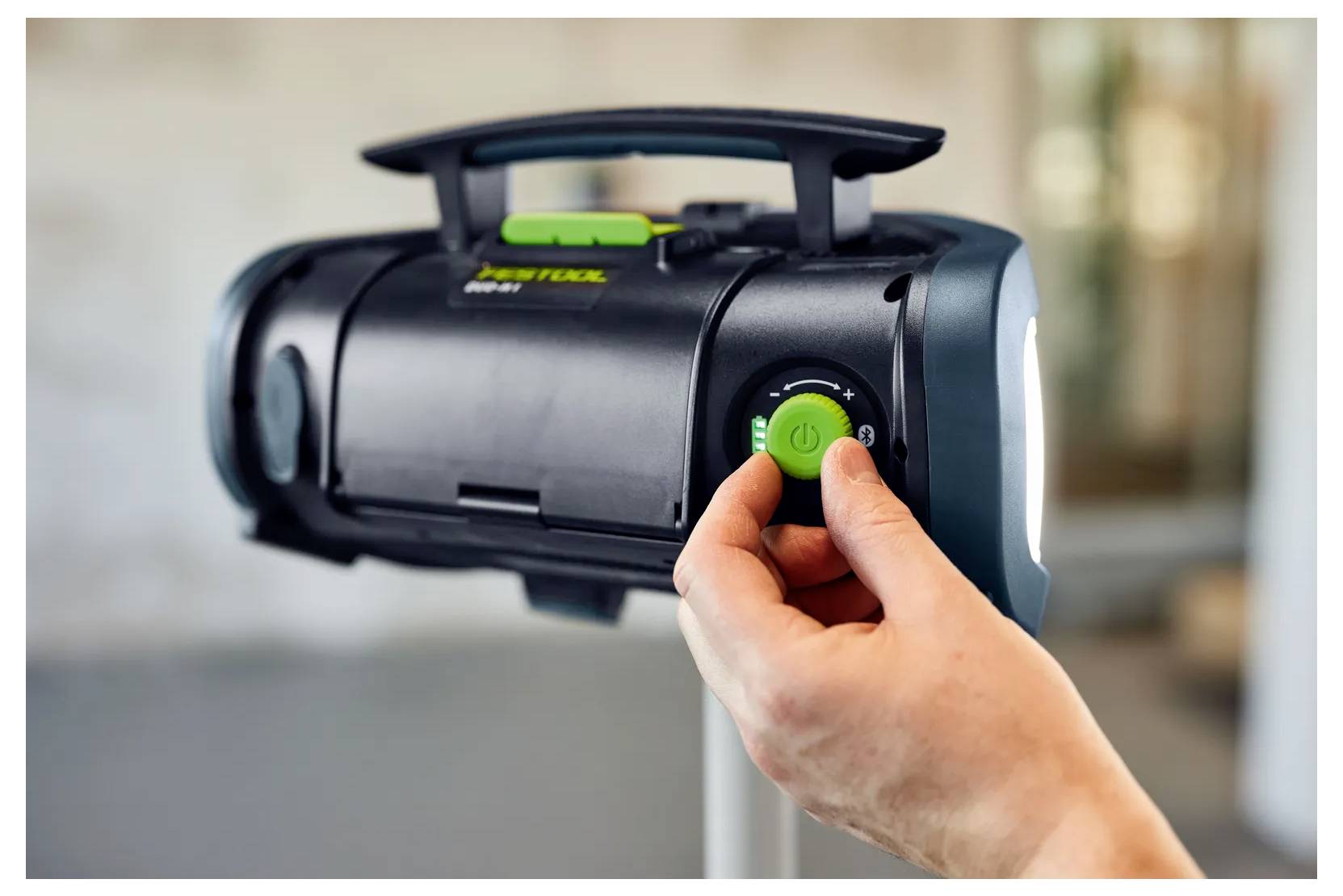 Hand die de aan/uit-knop op een zwarte draagbare speaker met handvat en groene bedieningselementen omdraait.