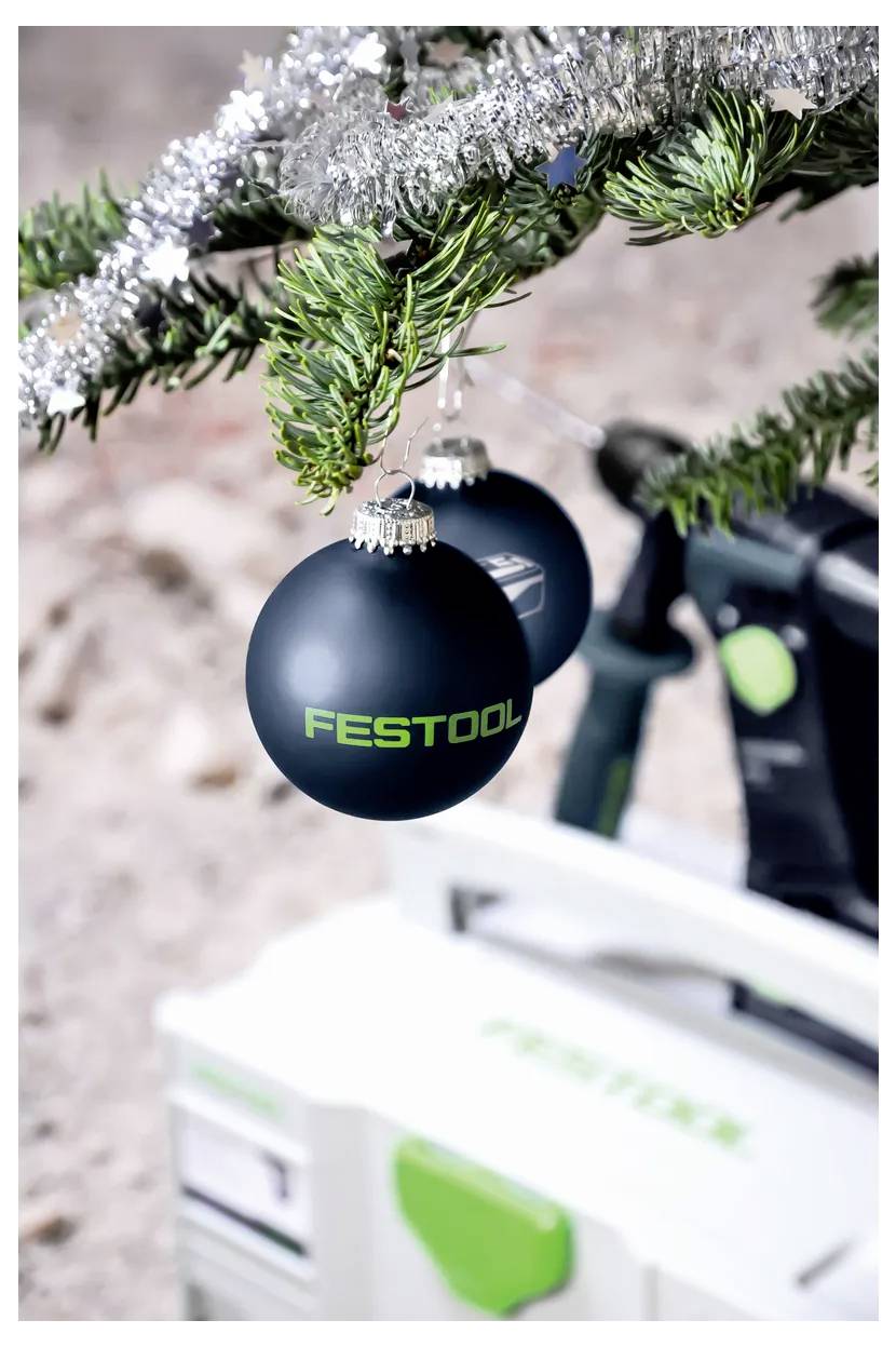 Zwart Festool ornament hangend aan een takje van een kerstboom, met een vervaagde Festool gereedschapskist op de achtergrond.