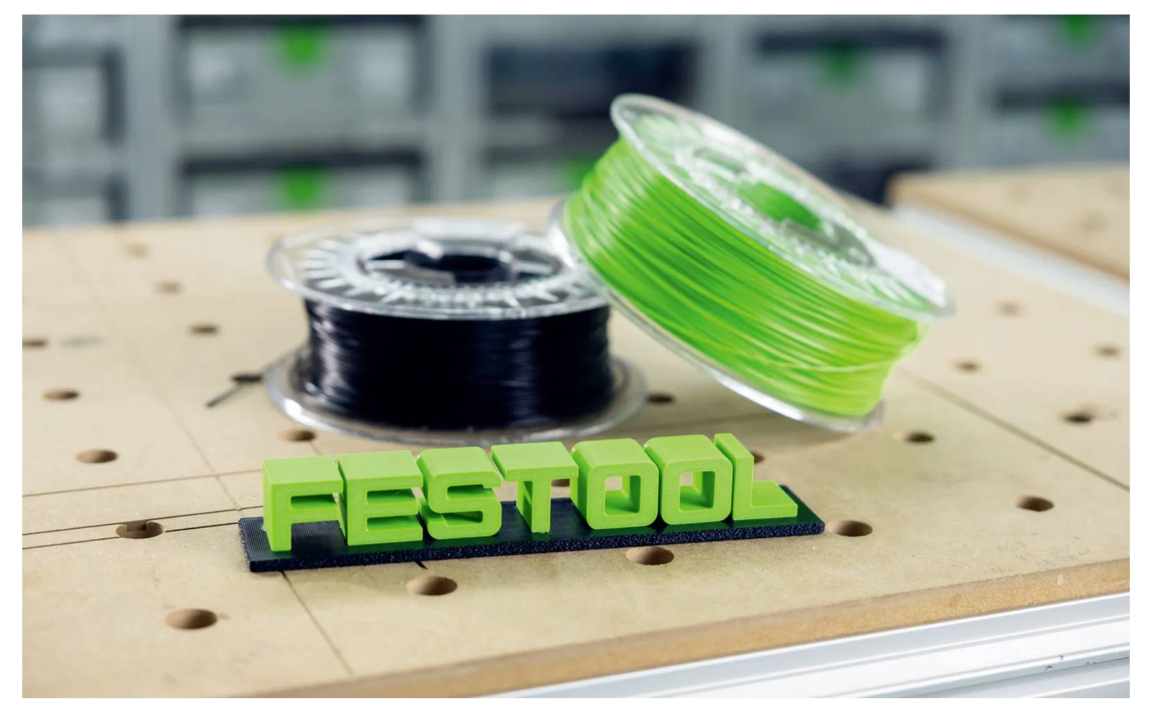 Groen 3D-geprinte 'FESTOOL'-logo op een werkbank, met zwarte en neongroene filamentspolen erachter in een werkplaatsinrichting.