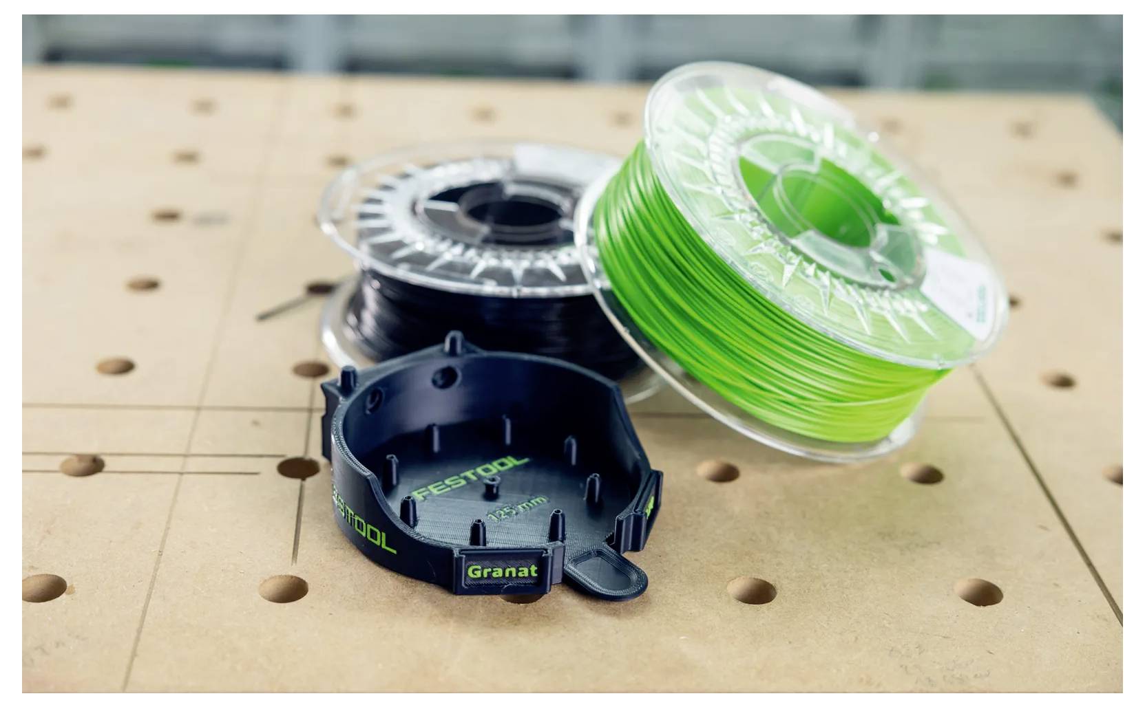 Drie 3D-printerspoelenmet filament, waaronder felgroen filament, op een geperforeerd werkoppervlak, met een klein zwart geprint onderdeel ervoor.