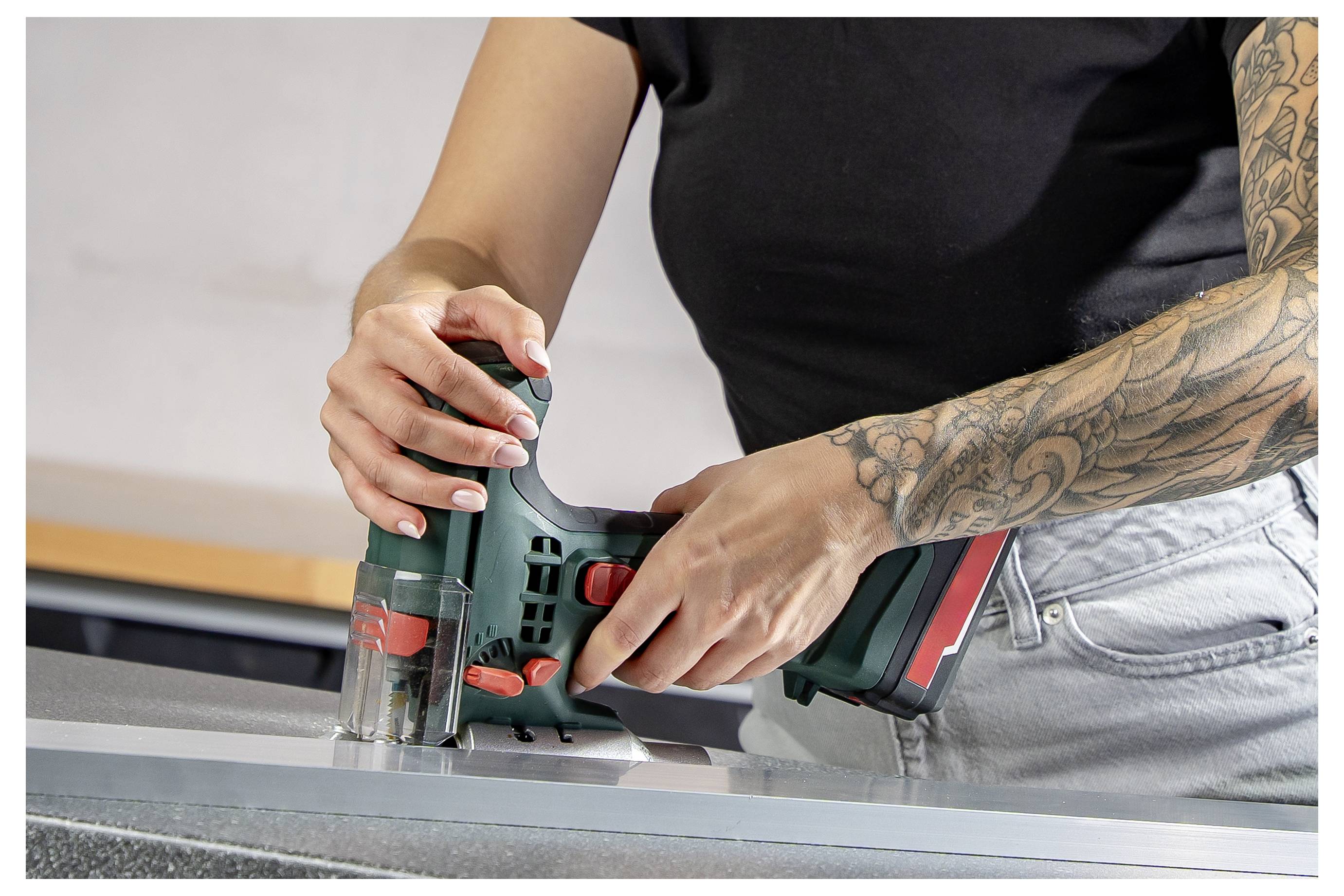 Een persoon die een draadloze figuurzaag gebruikt om een materiaaldeel op een werkbank door te zagen, met een getatoeëerde arm zichtbaar.