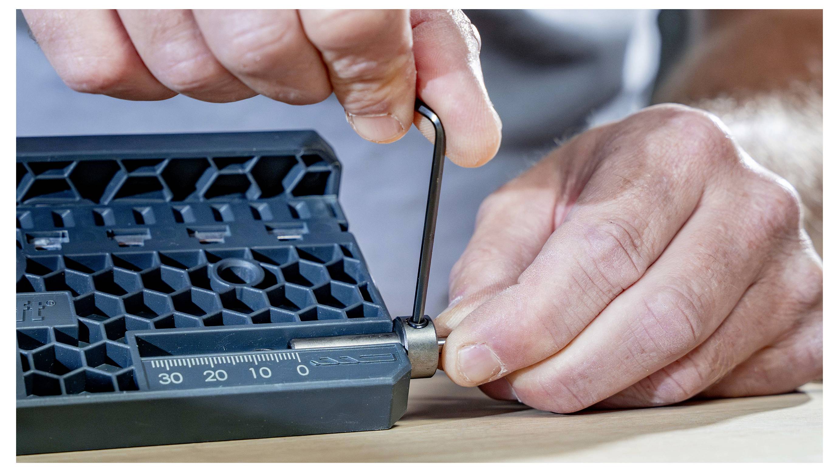 Handen die een inbussleutel gebruiken om een schroef aan te draaien op een zwart plastic zeshoekig-raster component met meetmarkeringen.