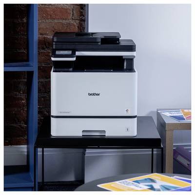 Witte Brother printer/kopieerapparaat op een zwart statief naast een bakstenen muur en een lichtgekleurde muur, met papieren op nabijgelegen tafels.