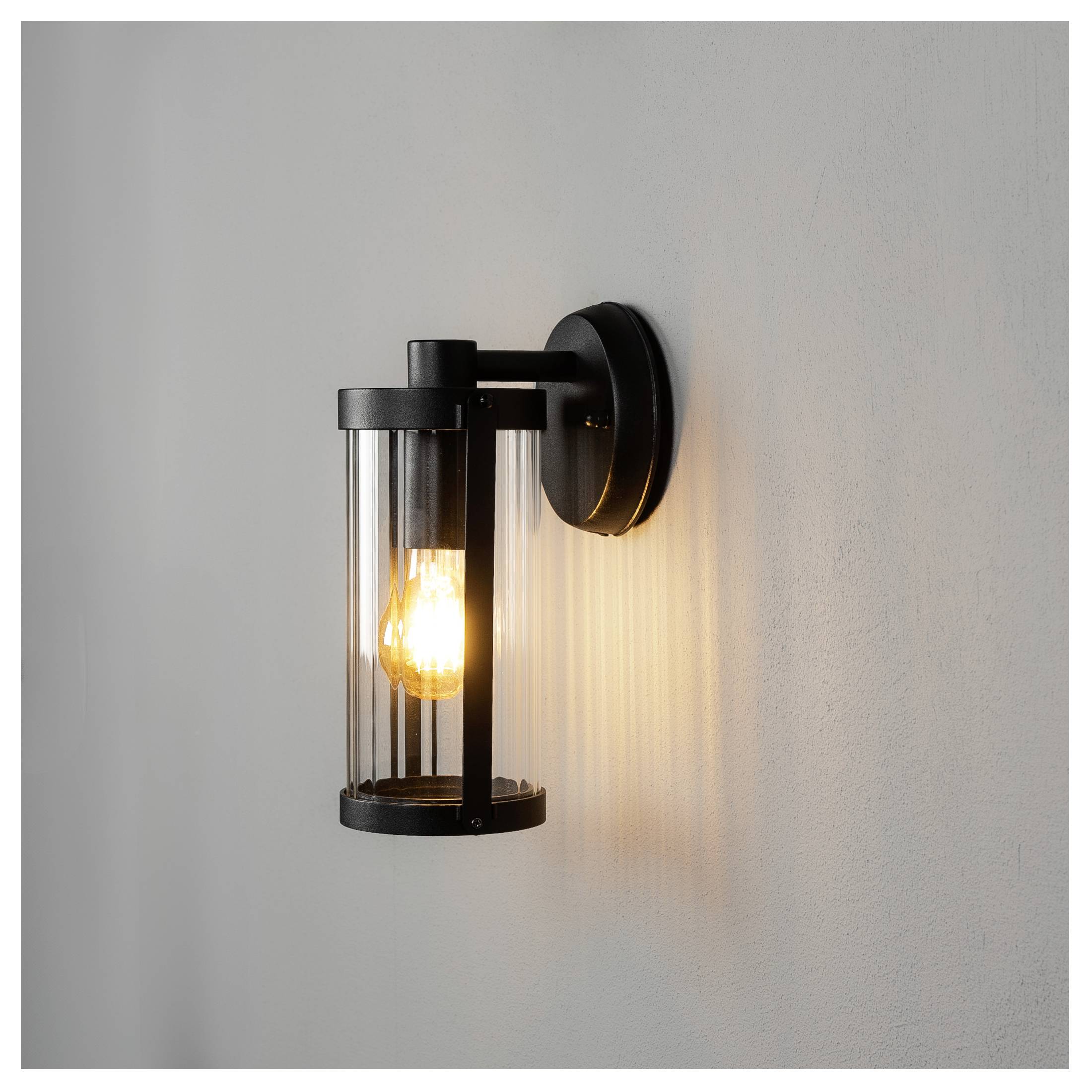 Zwarte cilindervormige wandlamp met zichtbare gloeiende lamp op een lichtgrijs oppervlak, die een warm verticaal lichtpatroon werpt.