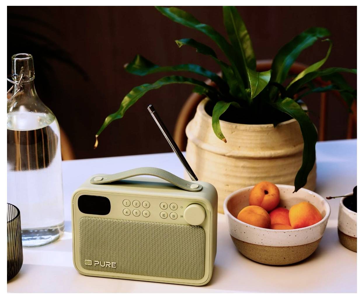 Een kleine beige draagbare radio met een handvat staat op een tafel naast een waterfles, een plant en een schaal met fruit.