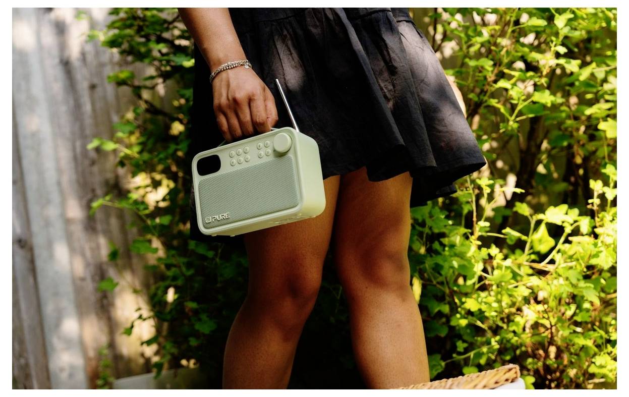 Een persoon in een zwarte jurk die een kleine mintgroene draagbare speaker buiten vasthoudt naast een bladrijke muur.
