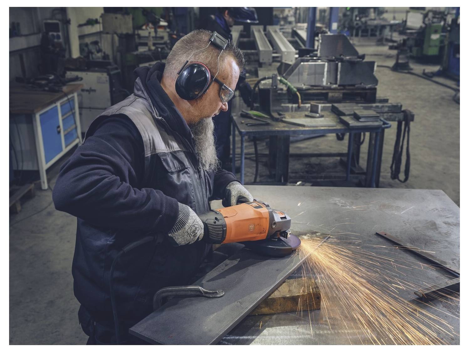 Een werknemer die gehoorbescherming draagt, slijpt een metalen plaat met een haakslipmachine, waarbij vonken door een industriële werkplaats vliegen.