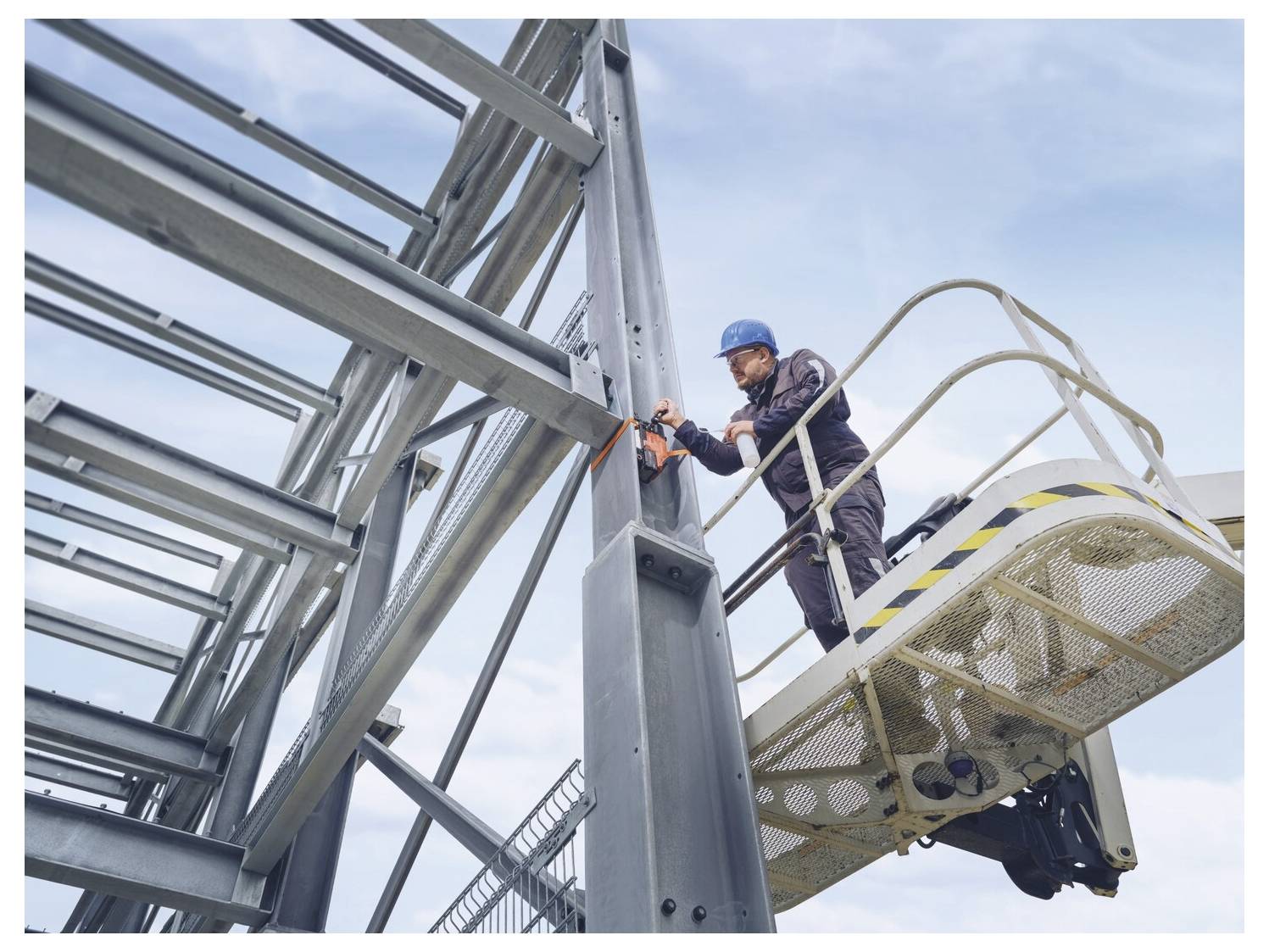 Bouwvakker in een veiligheidshelm staat op een verhoogd platform en bevestigt een stalen balk aan een metalen frame tegen een blauwe lucht.