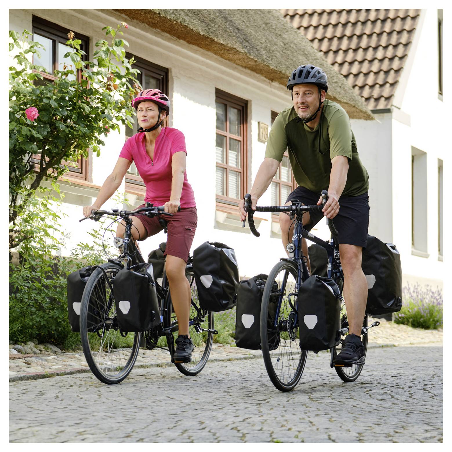 Twee fietsers die helmen dragen, rijden op toerfiets met tassen op een kasseienstraat naast huizen.