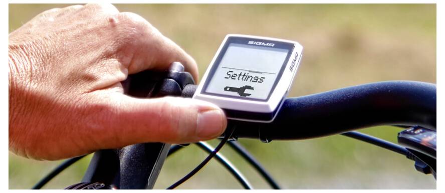 Hand die een fietscomputer op het stuur aanpast, waarbij 'Instellingen' op het scherm wordt weergegeven.