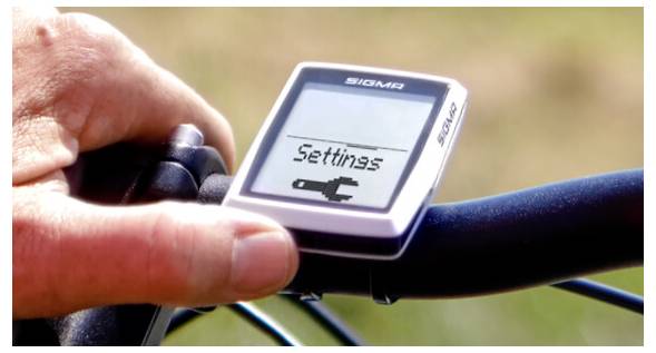 Een hand drukt op een knop van een Sigma fietscomputer terwijl het scherm 'Instellingen' toont.