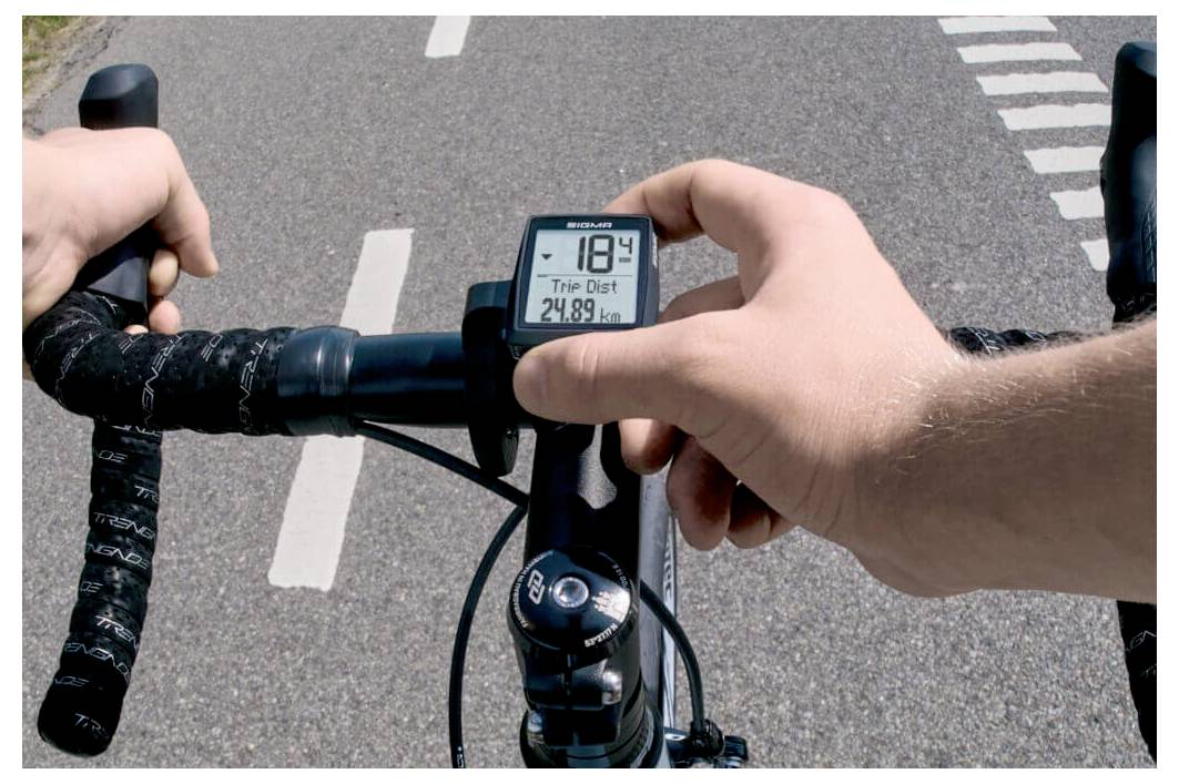 Handen van de fietser op het stuur terwijl hij op een weg rijdt, met een fietscomputer die een snelheid van 18,4 en een ritafstand van 24,89 km aangeeft.