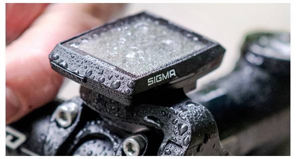 Close-up van een natte Sigma fietsmeter gemonteerd op een fietsstuur, met waterdruppels op het apparaat en het fietsframe.