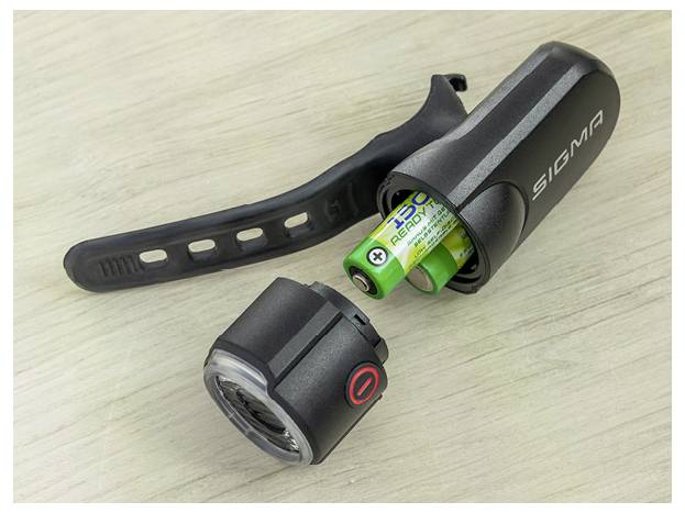 Gedemonteerde Sigma-fietslamp met verwijderde batterijen, waarop de lampbehuizing, lensdop en zwarte montageband op een licht houten oppervlak zichtbaar zijn.