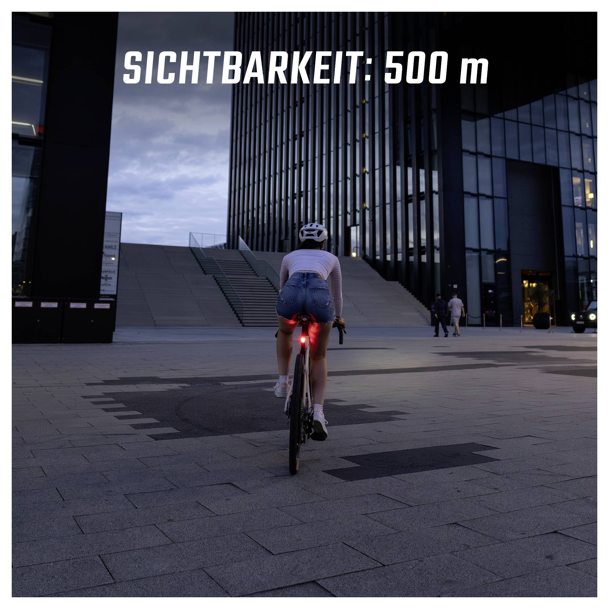 Achteraanzicht van een fietser in de schemering voor moderne gebouwen; rood achterlicht brandt, tekst: 'ZICHTBAARHEID: 500 m'.