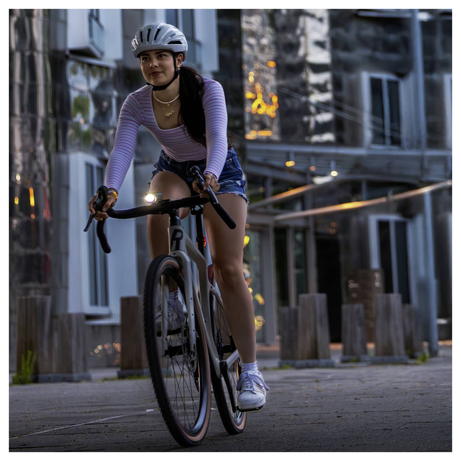 Jonge vrouw rijdt met racefiets op een stedelijk voetpad in de schemering; zij draagt een helm, gestreept bovenstuk en shorts.