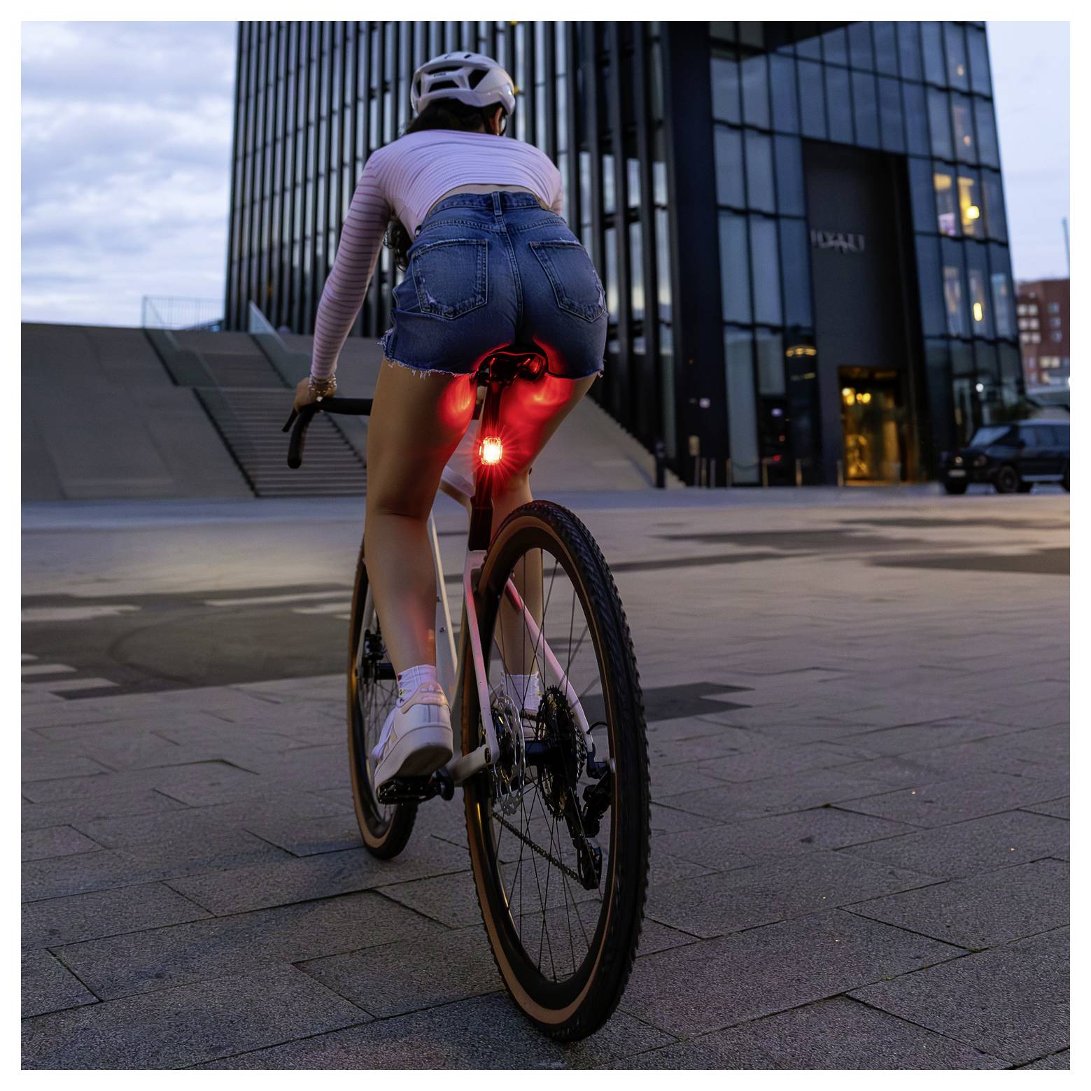 Wielrenster met helm rijdt bij schemering voor een modern kantoorgebouw; rood achterlicht op de fiets brandt duidelijk.