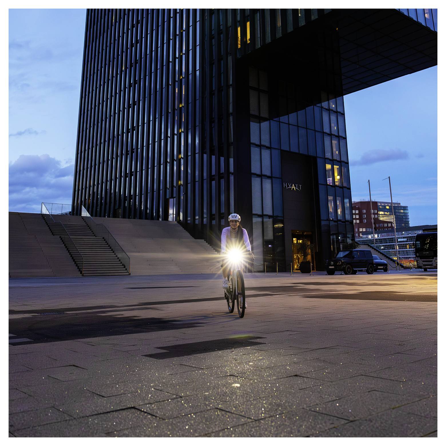 Fietser met ingeschakeld voorlicht rijdt tijdens het schemeren voor een modern wolkenkrabber op een leeg plein.