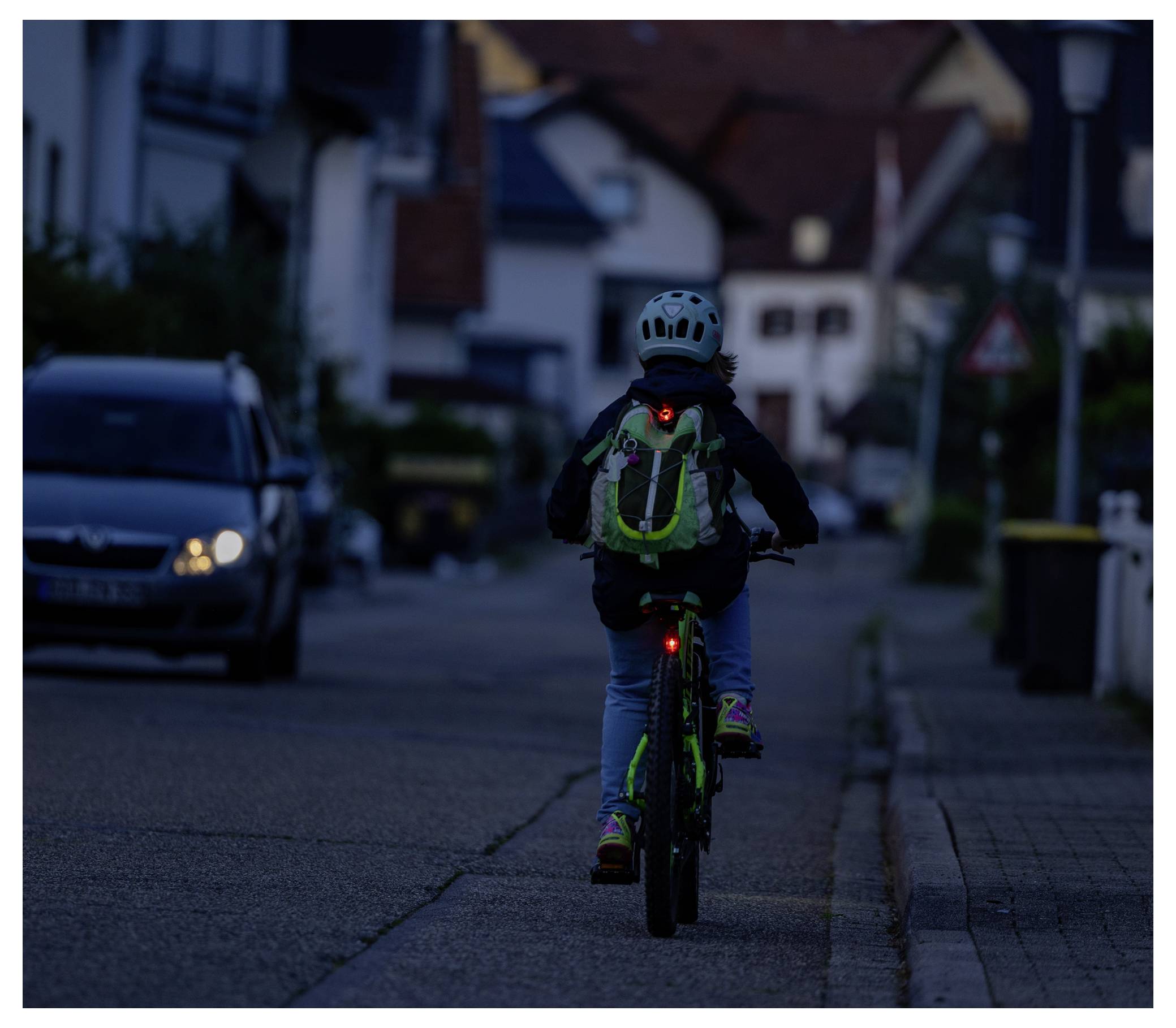 Kind rijdt op een fiets weg over een donkere woonstraat bij schemering, draagt een helm en rugzak met een zichtbaar rood achterlicht.