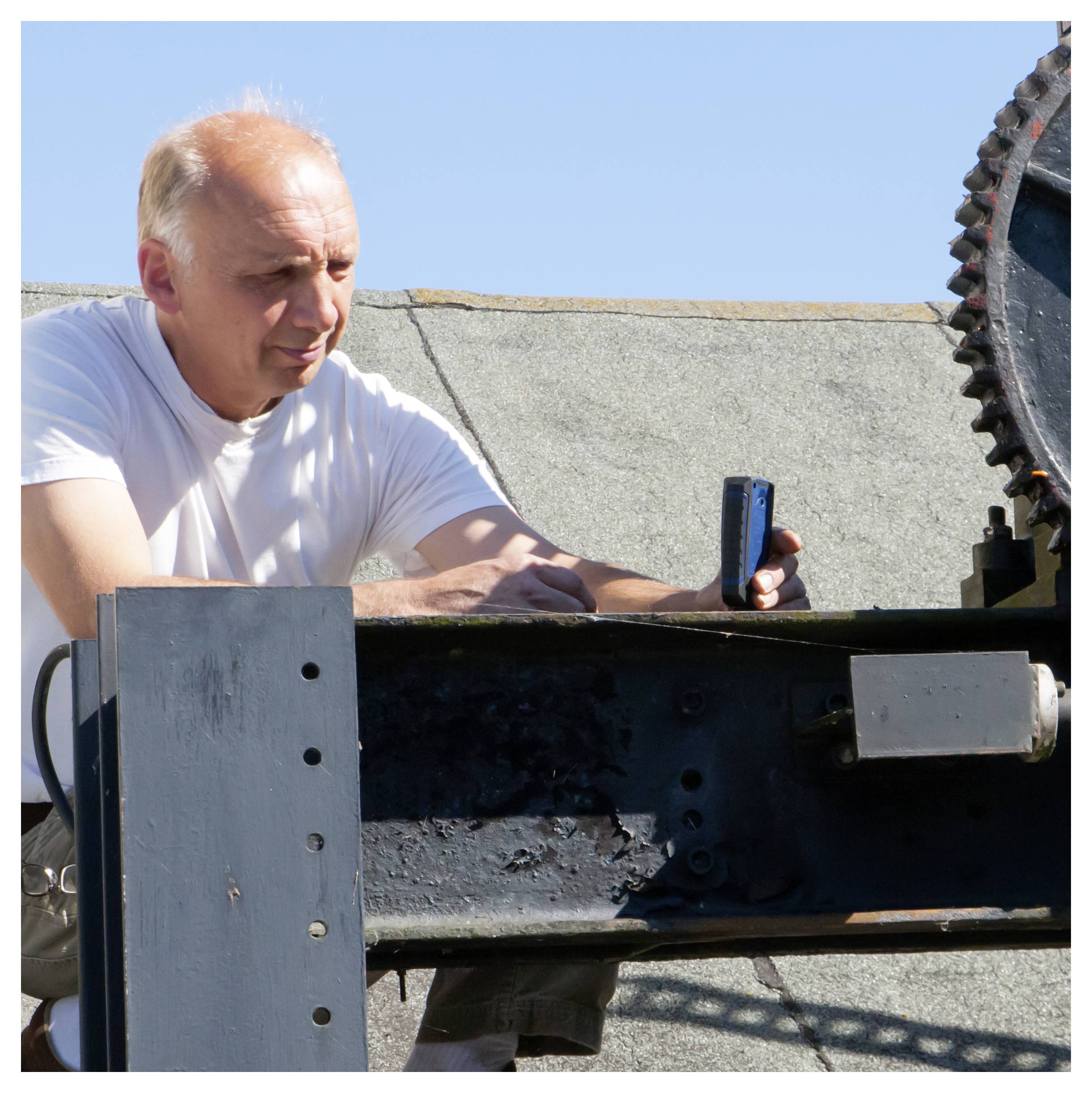 Oudere man in een wit T-shirt werkt buiten met een grote metalen zaag of snijmachine, houdt een gereedschap vast en inspecteert de opstelling.