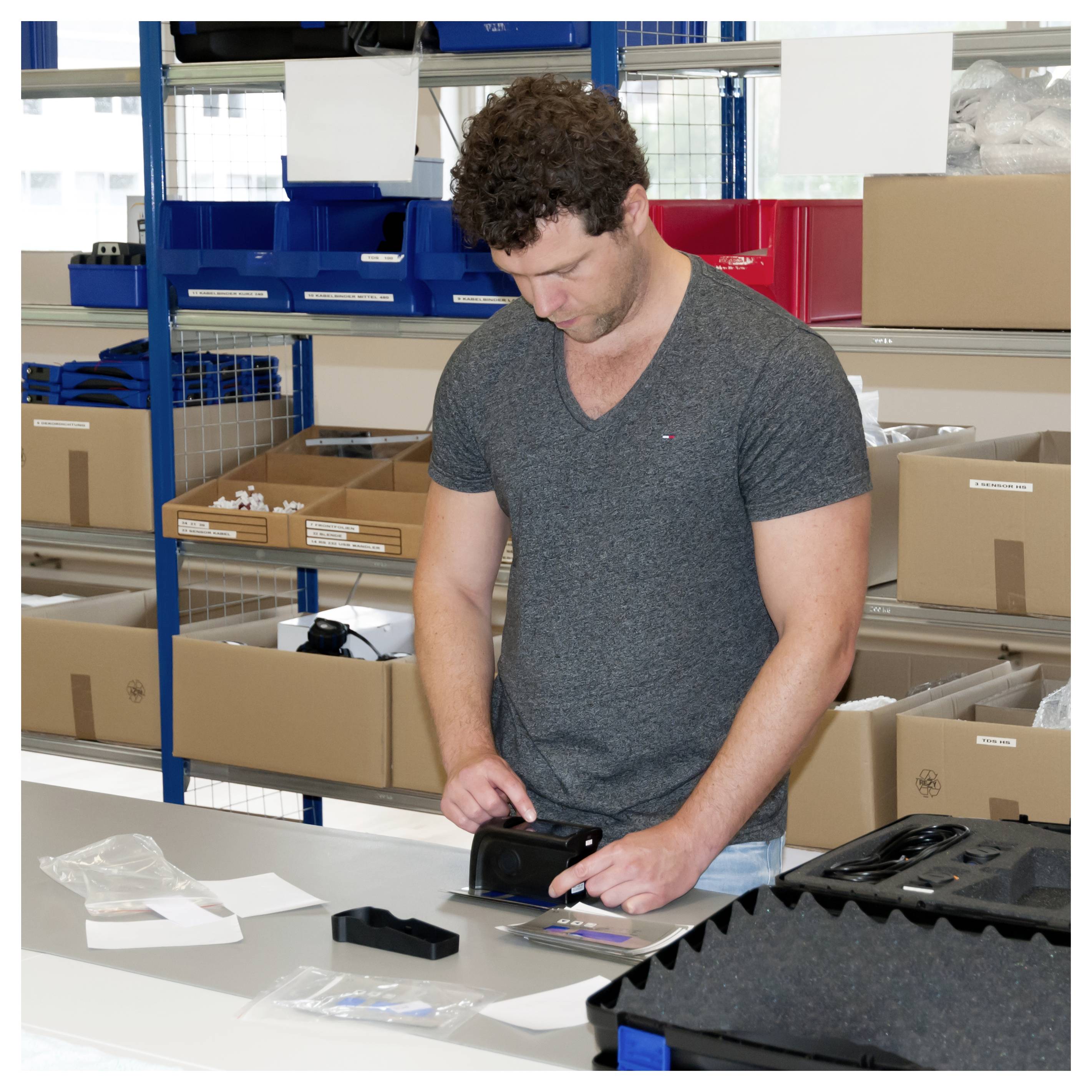 Man die een klein zwart apparaat op een werkttafel in een magazijn inspecteert, met verpakte voorraad en opslagrekken achter hem.