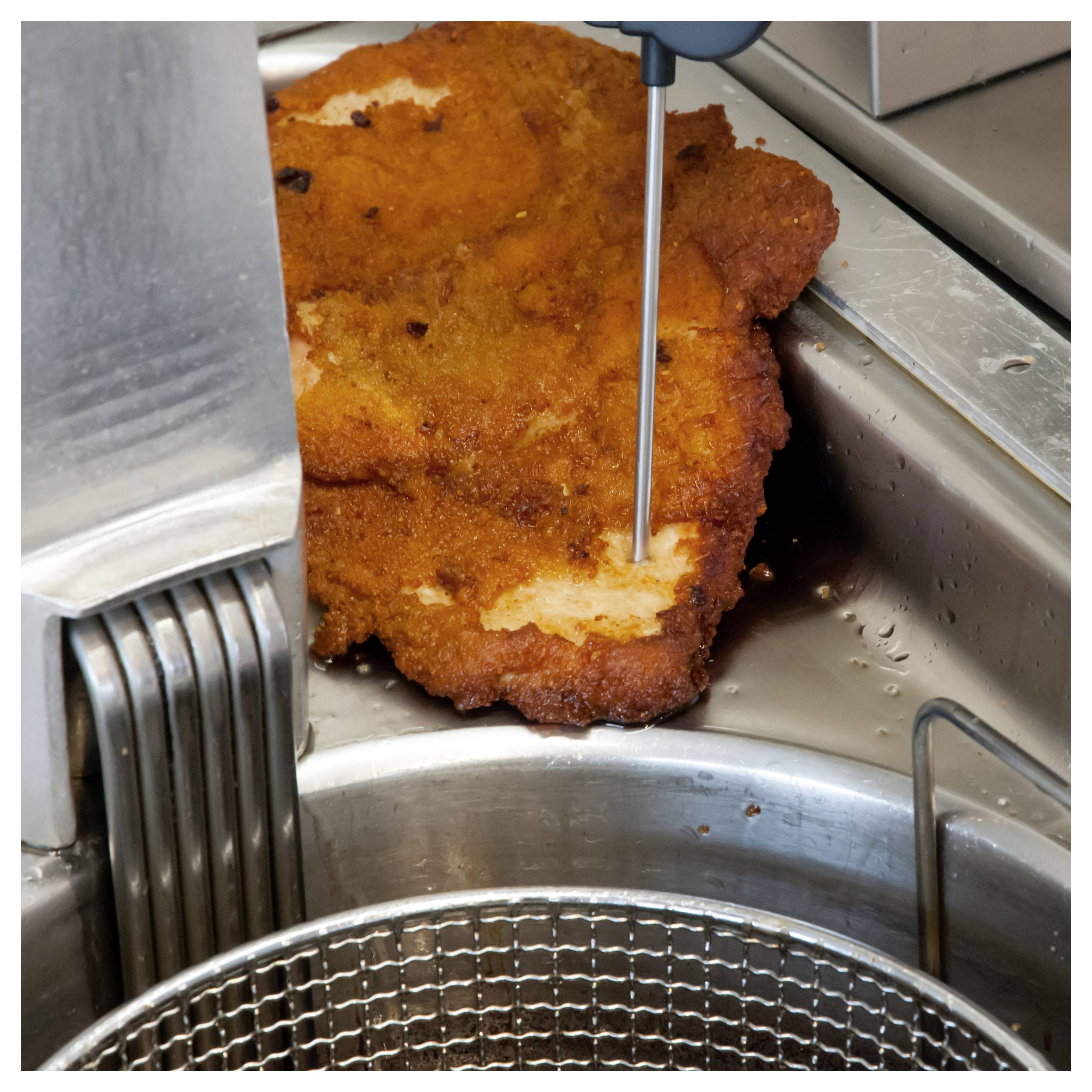 Een paneerbesteck wordt doorboord door een metalen thermometerstift nabij een friteuse, waarbij de gaarmaaktemperatuur wordt gecontroleerd.