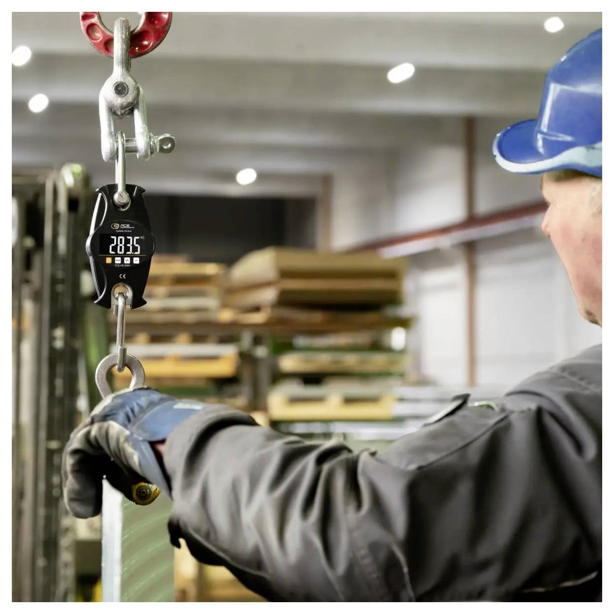 Arbeider met een veiligheidshelm hanteert een hangende last met een digitale kraanweegschaal die 2835 aangeeft in een industrieel magazijn.