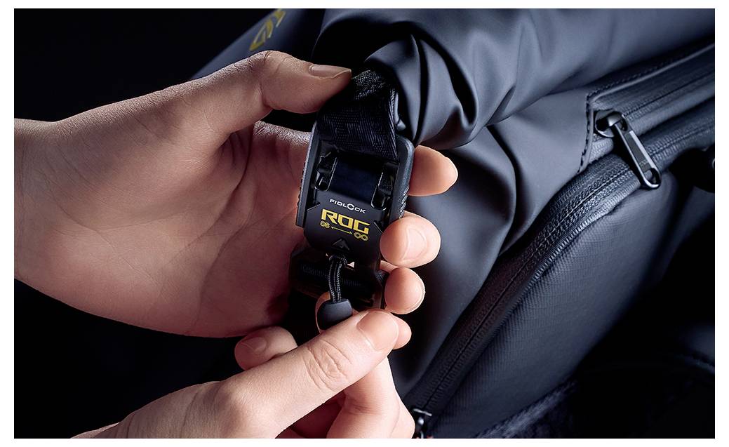 Handen die een zwarte Rog CPX-clip aan een zwarte rugzakband naast een geritste zak bevestigen, wat aantoont hoe een accessoire aan een tas wordt bevestigd.