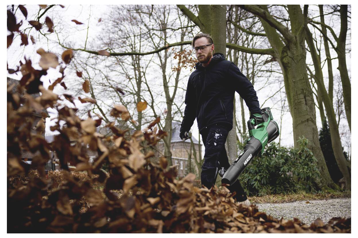 Man die buiten in een park loopt, een handhelds bladblazer gebruikt om een stapel droge herfstbladeren van een gravelpad schoon te maken.