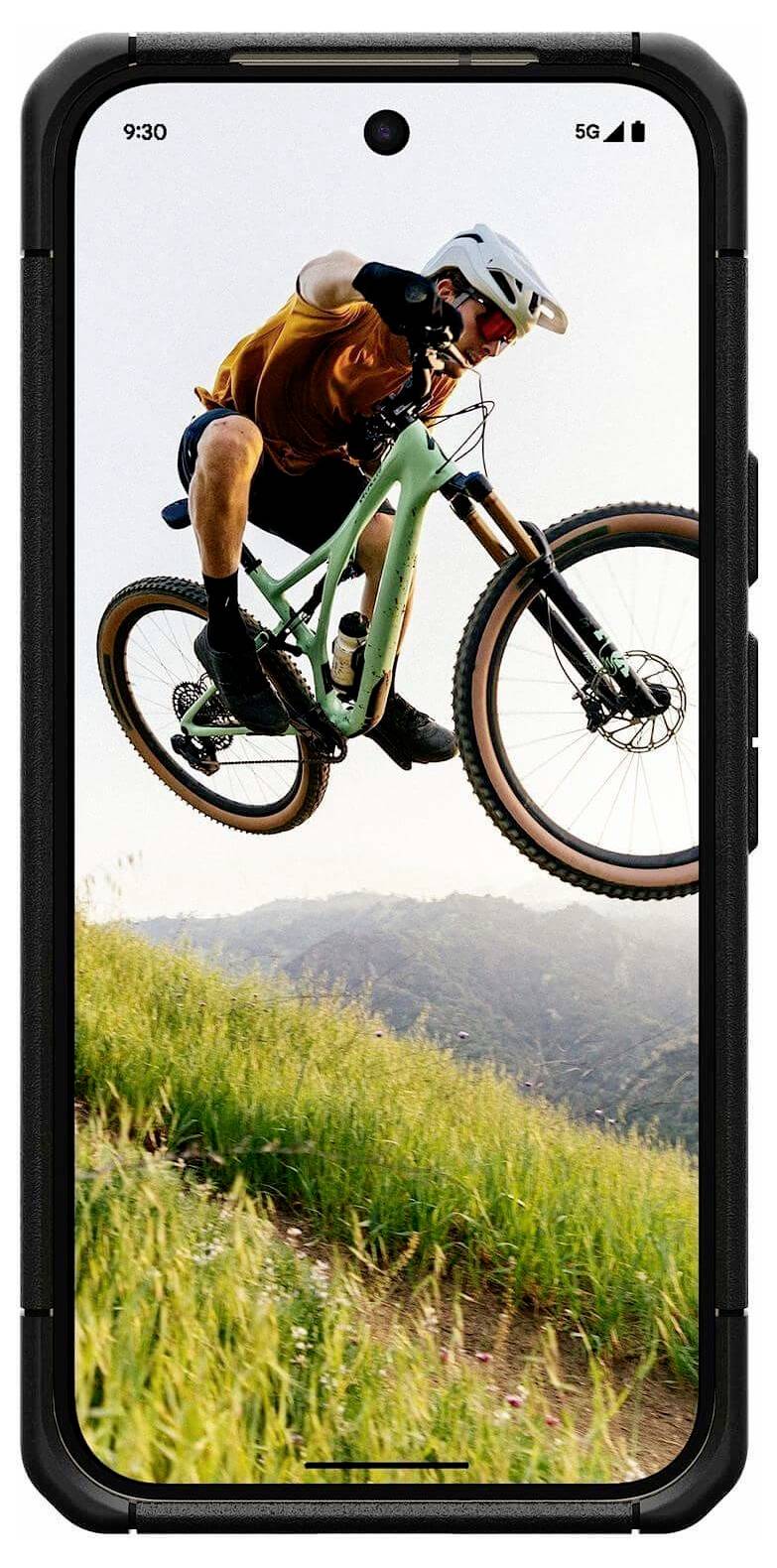Smartphonescherm met een mountainbiker in de lucht tijdens een sprong over een grashelling, met heuvels op de achtergrond.