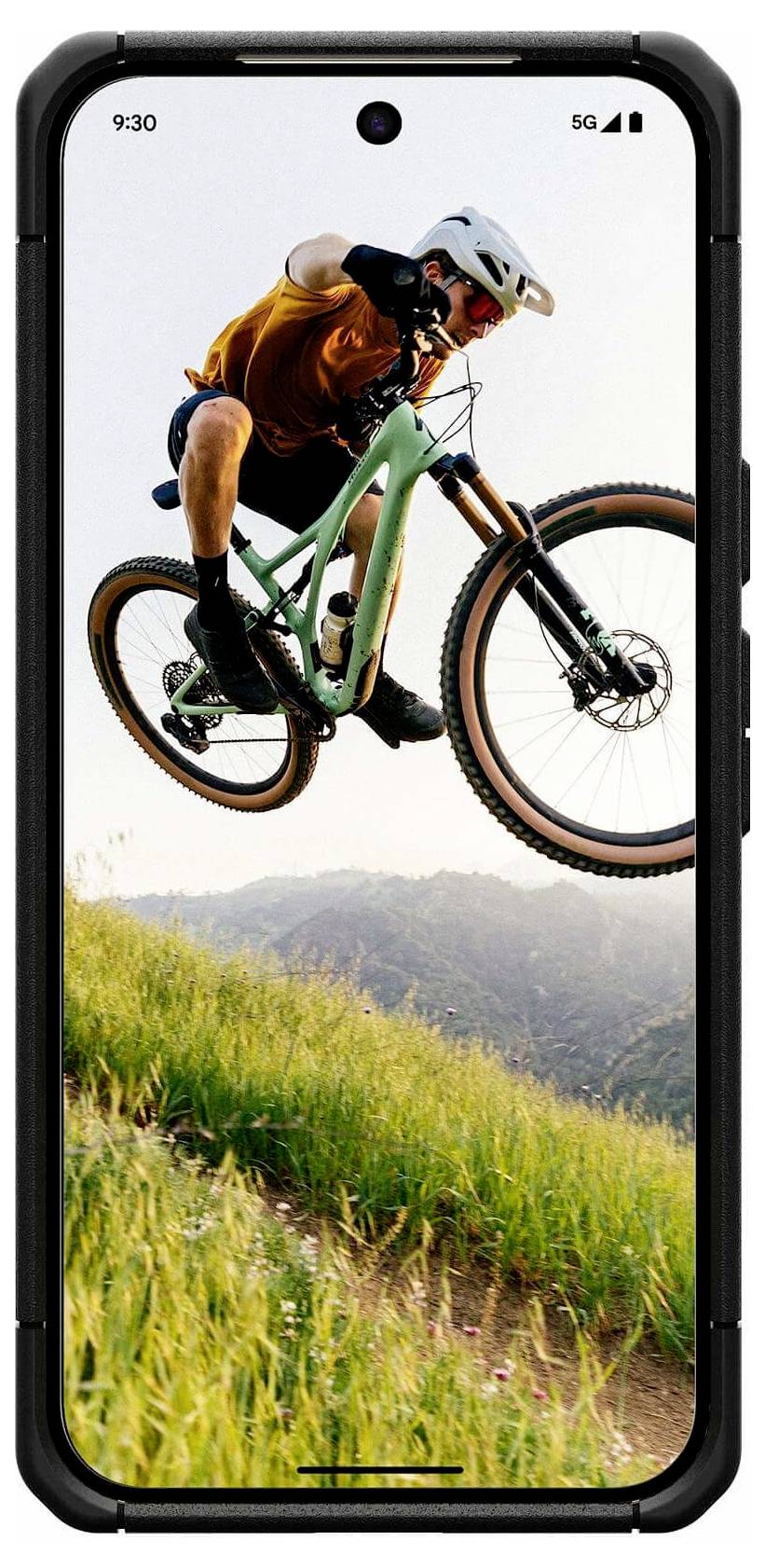 Een mountainbiker midden in een sprong op een trail, in de lucht boven grasbedekte heuvels bij zonsondergang, weergegeven op een smartphonescherm.