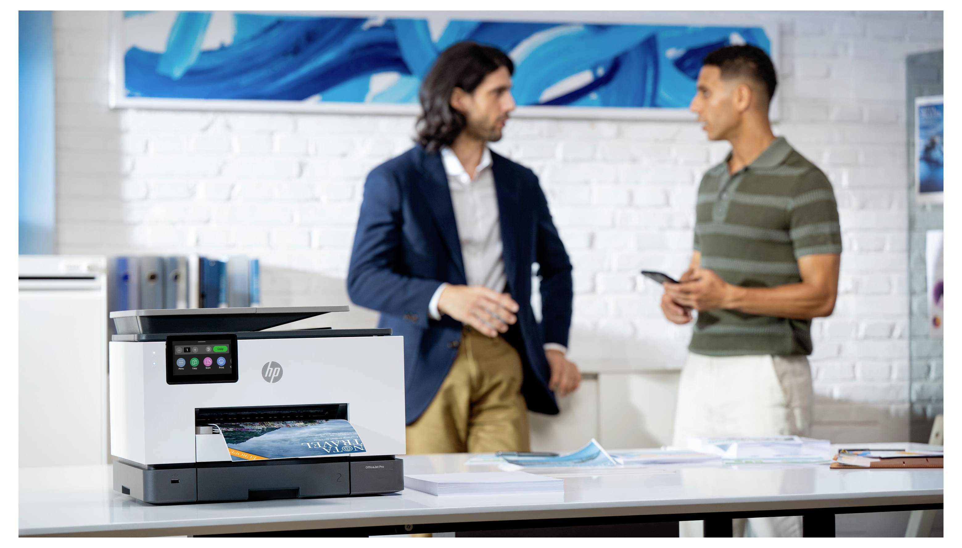 Twee mannen in gesprek in een modern kantoor naast een HP-printer op een bureau, met documenten en een laptop op de voorgrond.