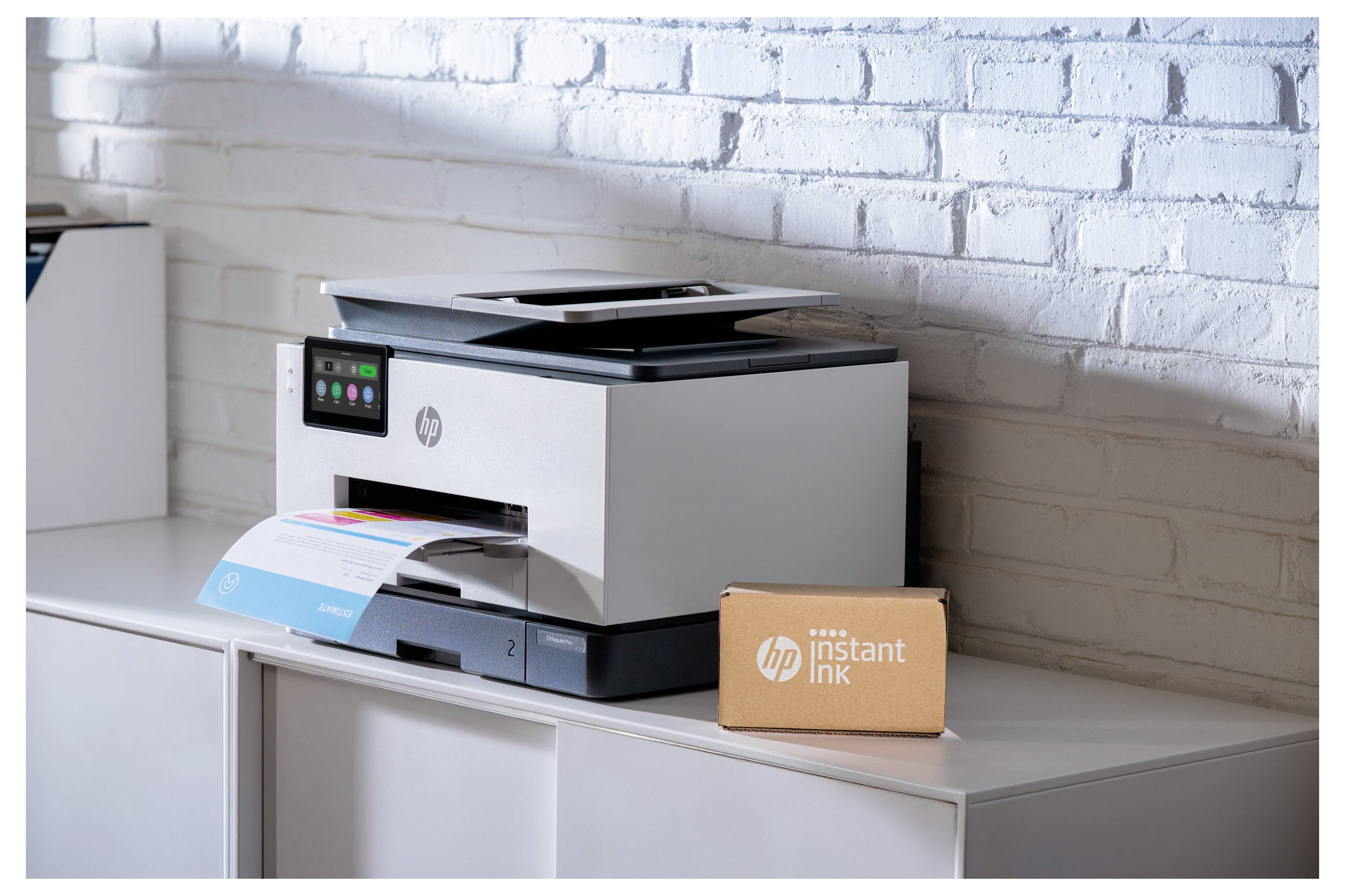 HP-printer op een kast naast een 'instant ink'-doos, met een afgedrukt blad dat eruit komt tegen een witte bakstenen muur.