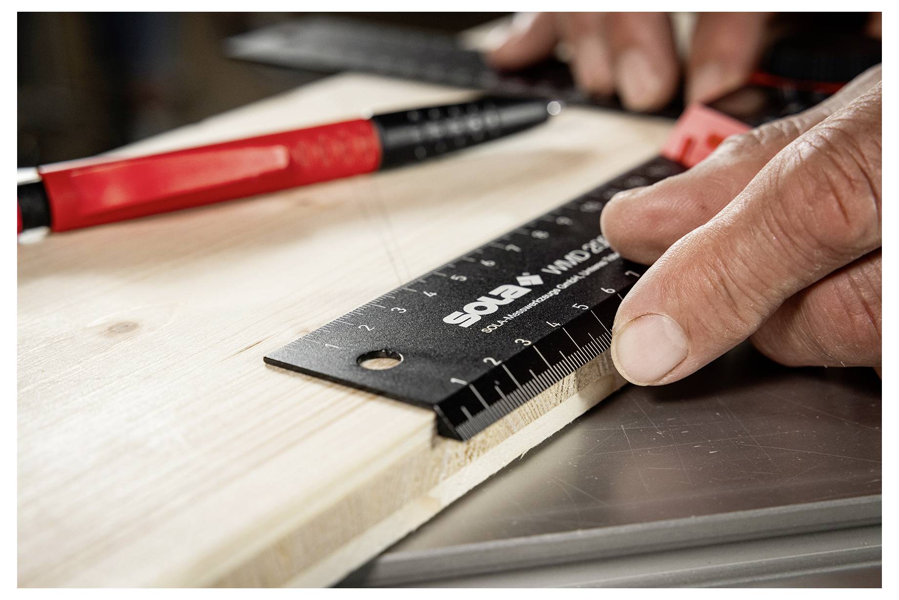 Closeup van een persoon die een houten plank meet met een zwarte liniaal terwijl een rood handgereedschap in de buurt op een werkblad rust.