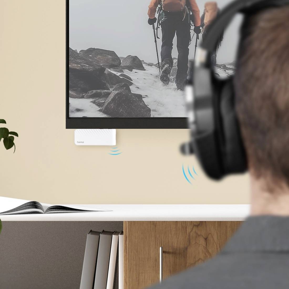 Persoon met koptelefoon luistert naar een aan de muur bevestigd scherm dat een wandelaar in sneeuwrijk terrein toont, met pictogrammen van draadloze audiosignalen die geluidstransmissie aangeven.