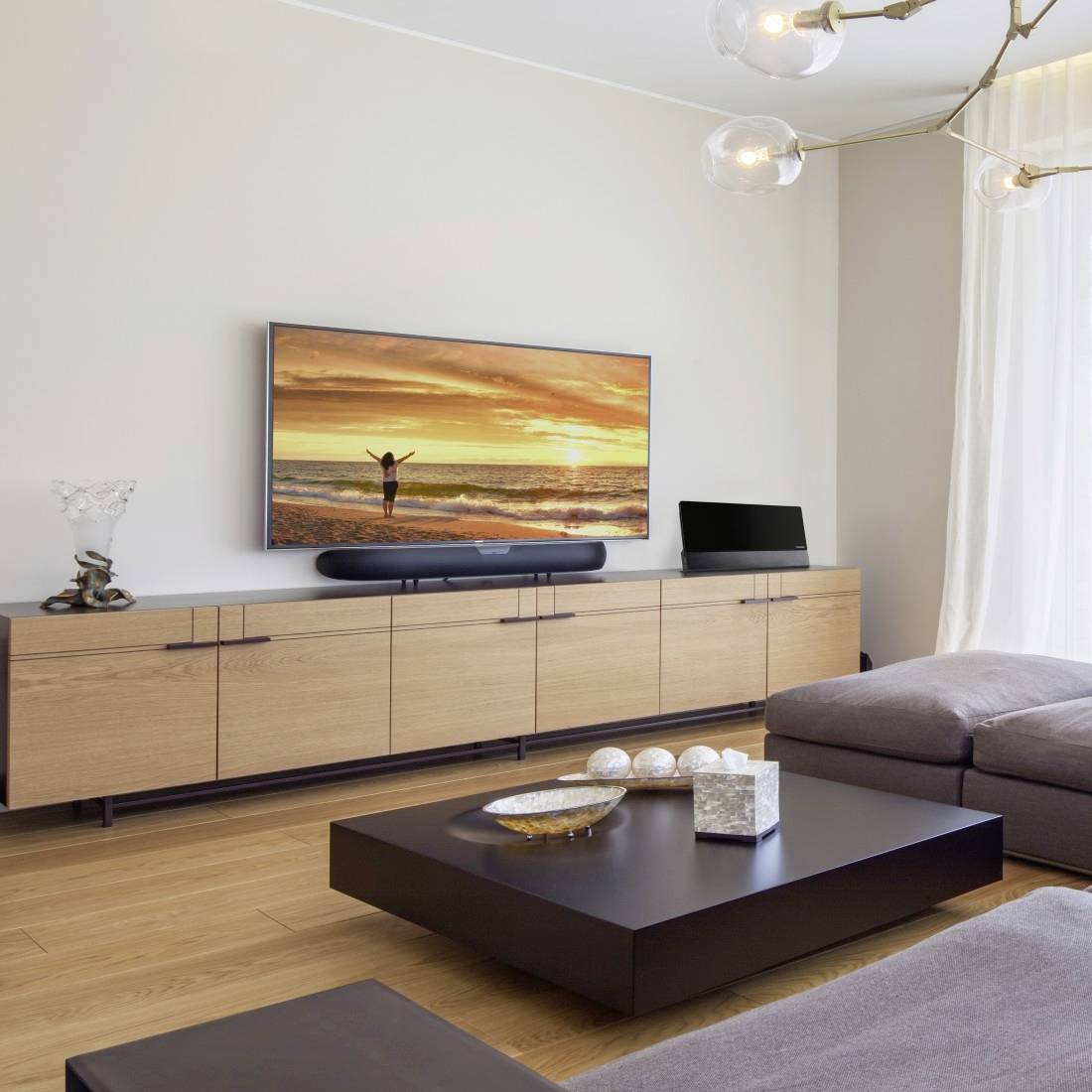 Moderne woonkamer met een aan de muur bevestigde televisie die een zonsondergangtafereel op het strand weergeeft, een laag houten kastje, een donkere salontafel, een hoekslaapbank en hangende lampen.