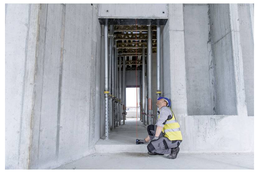 Bouwvakker in een veiligheidshelm en veiligheidsvest knielt onder een betonnen constructie en inspecteert een hangende schietlood in een bouwframe.