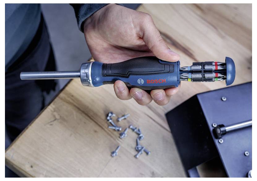 Hand die een Bosch-schroevendraaier met uitwisselbare bits vasthoudt boven een houten werkoppervlak, met verspreid liggende schroeven in de buurt.